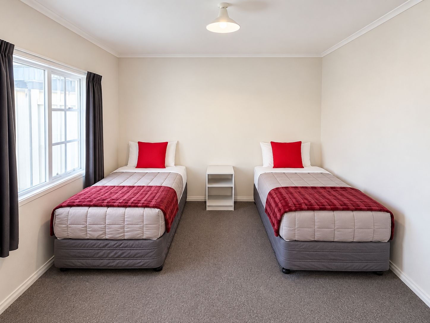A twin room with two beds, each with a red pillow, a white nightstand, and a window with curtains.