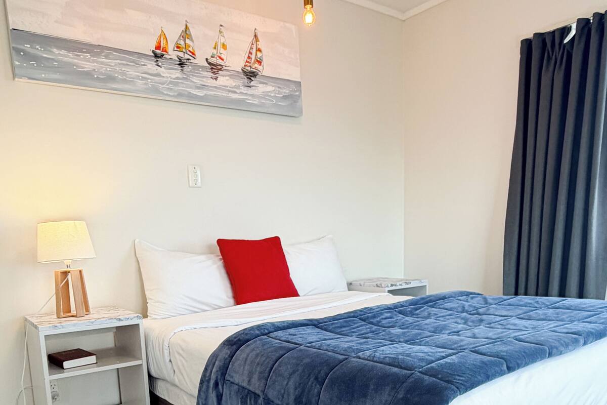 Ein Schlafzimmer mit Bett, Nachttisch, Lampe, einem Gemälde und einem Fenster mit Vorhängen.