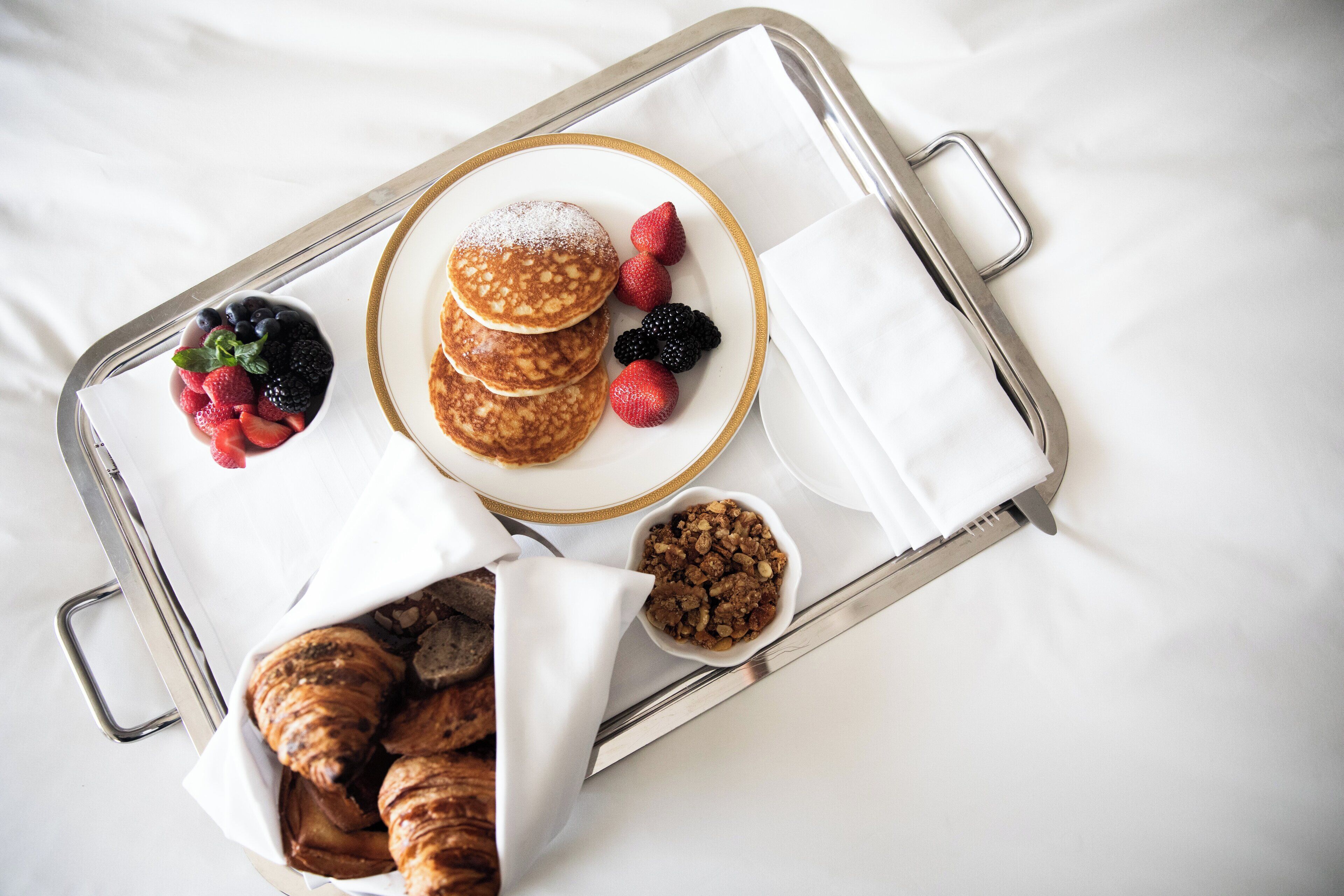 A tray with pancakes, berries, and croissants on a white surface.