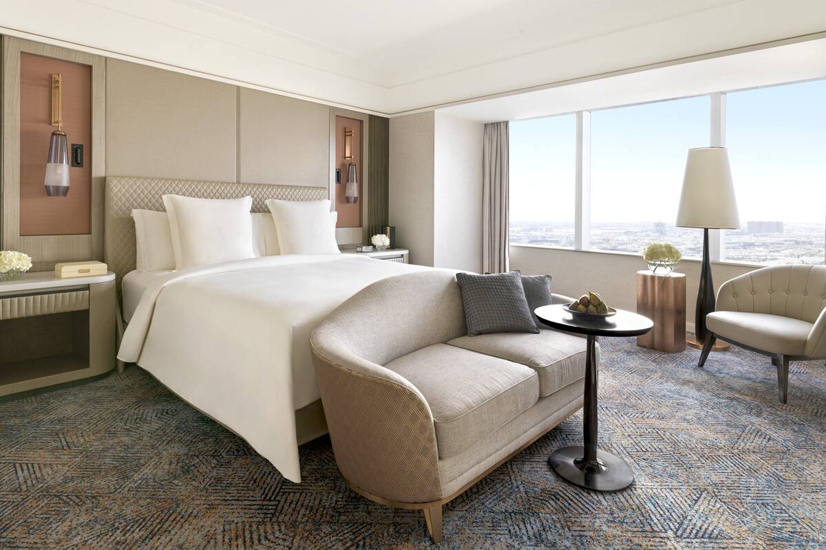 Habitación de hotel con una cama grande, un sofá, una mesita y una silla. Se ve una ventana con vistas a la ciudad.
