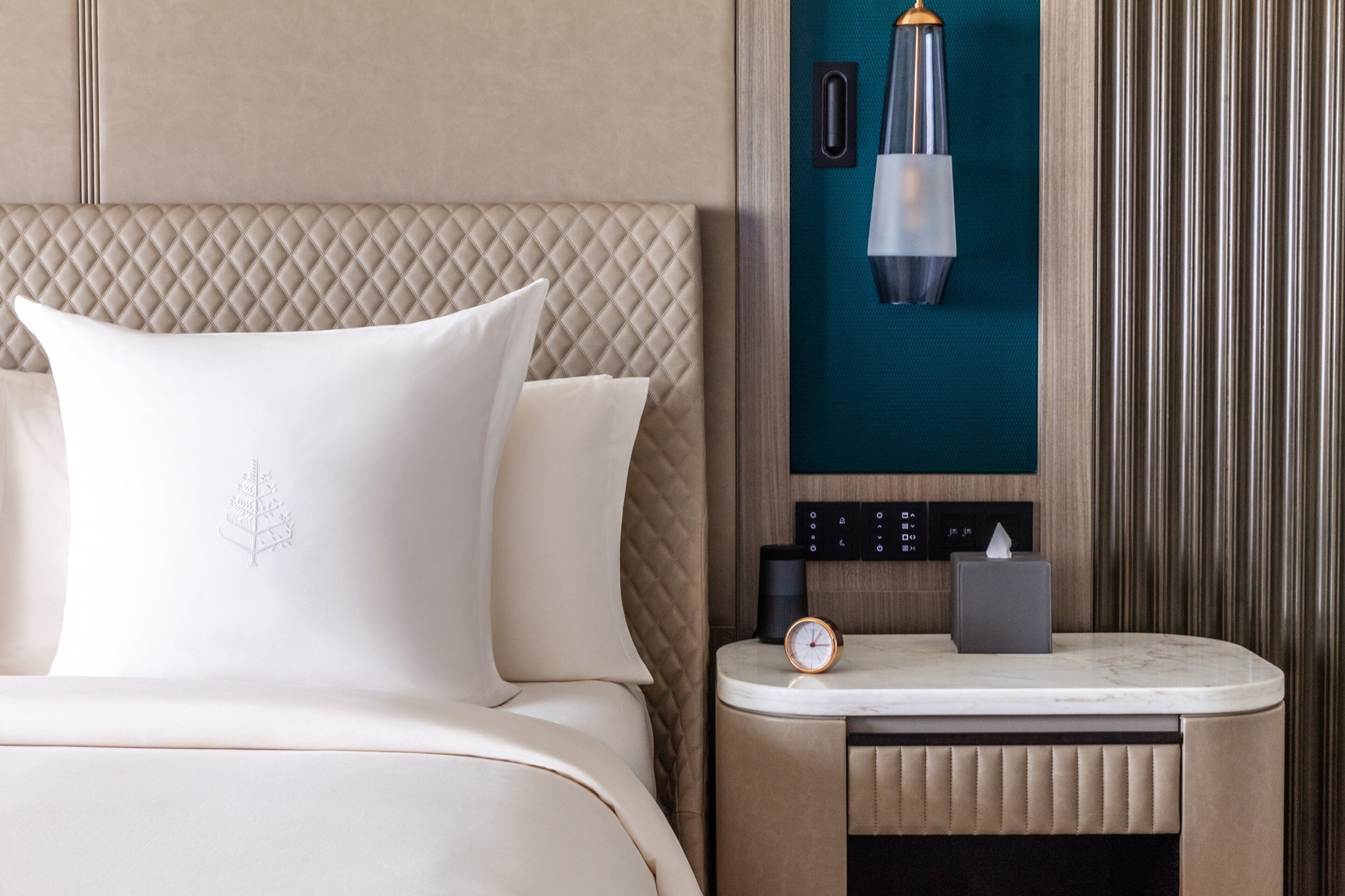 A hotel room with a bed, a nightstand, a clock, a lamp, and a wall-mounted control panel.
