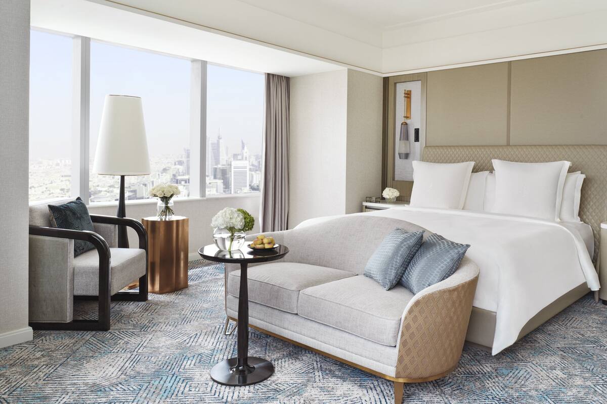 Habitación de hotel con una cama grande, un sofá, una mesita con flores, una lámpara y vistas a la ciudad.