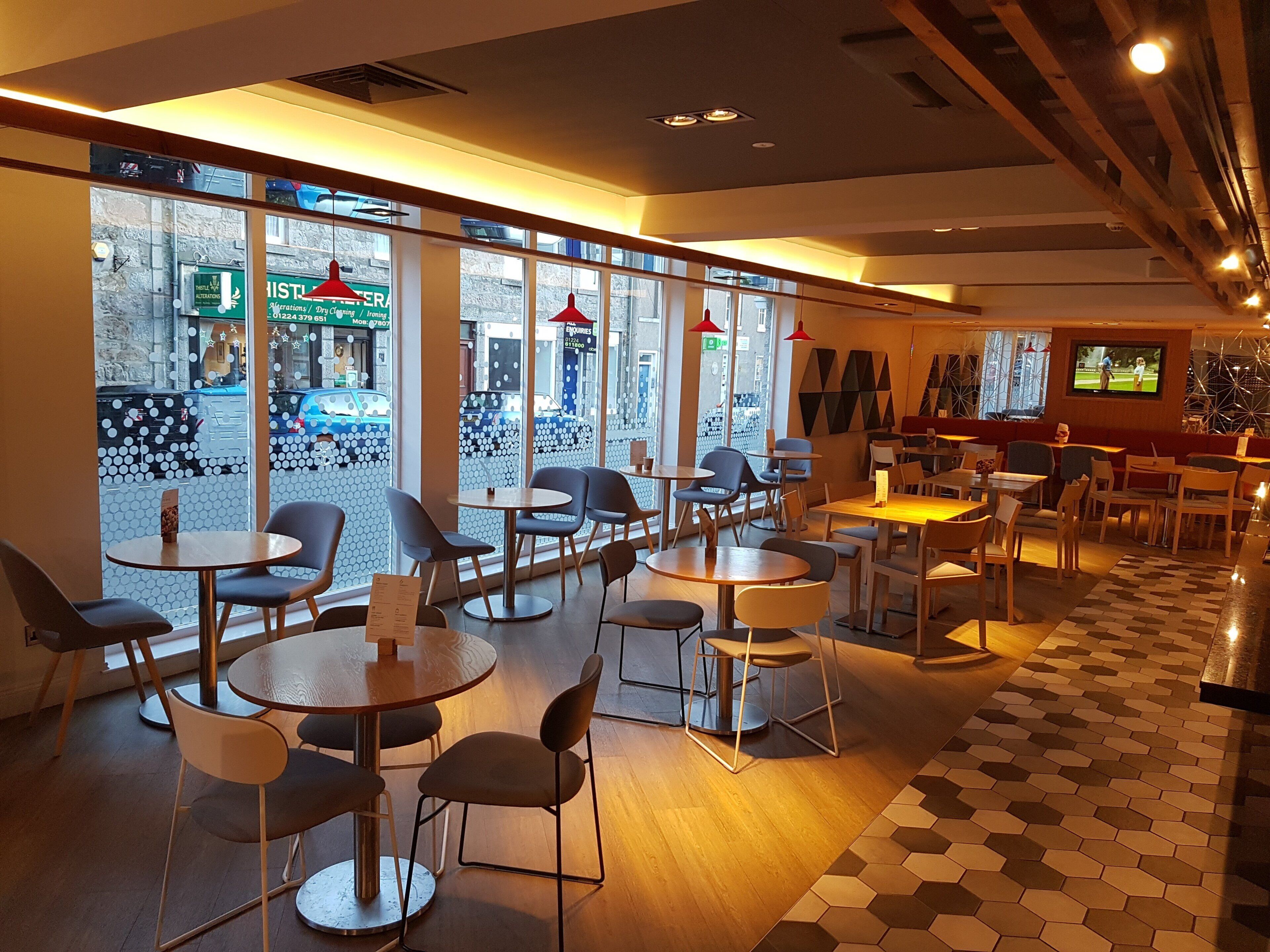 A restaurant with round tables and chairs, large windows with a view of the street, and a television screen on the wall.