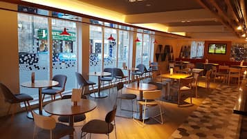 A restaurant with round tables and chairs, large windows with a view of the street, and a television screen on the wall.
