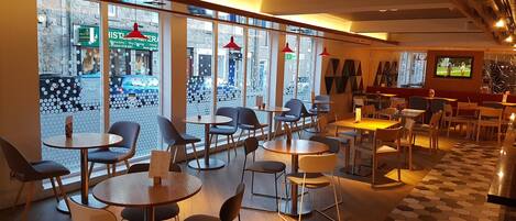 A restaurant with round tables and chairs, large windows with a view of the street, and a television screen on the wall.