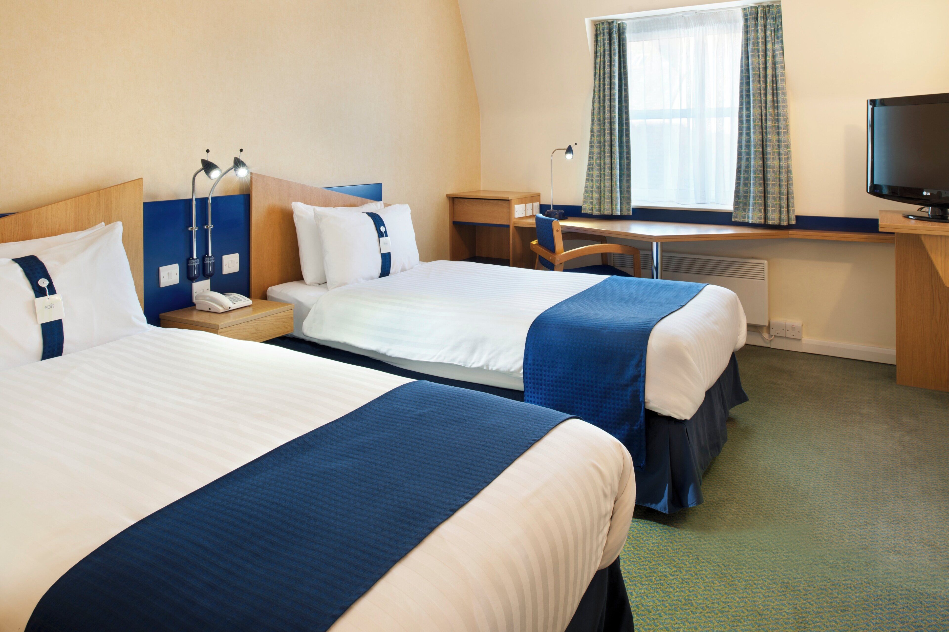 Une chambre d’hôtel avec deux lits, un bureau, une chaise, une télévision et une fenêtre avec des rideaux.