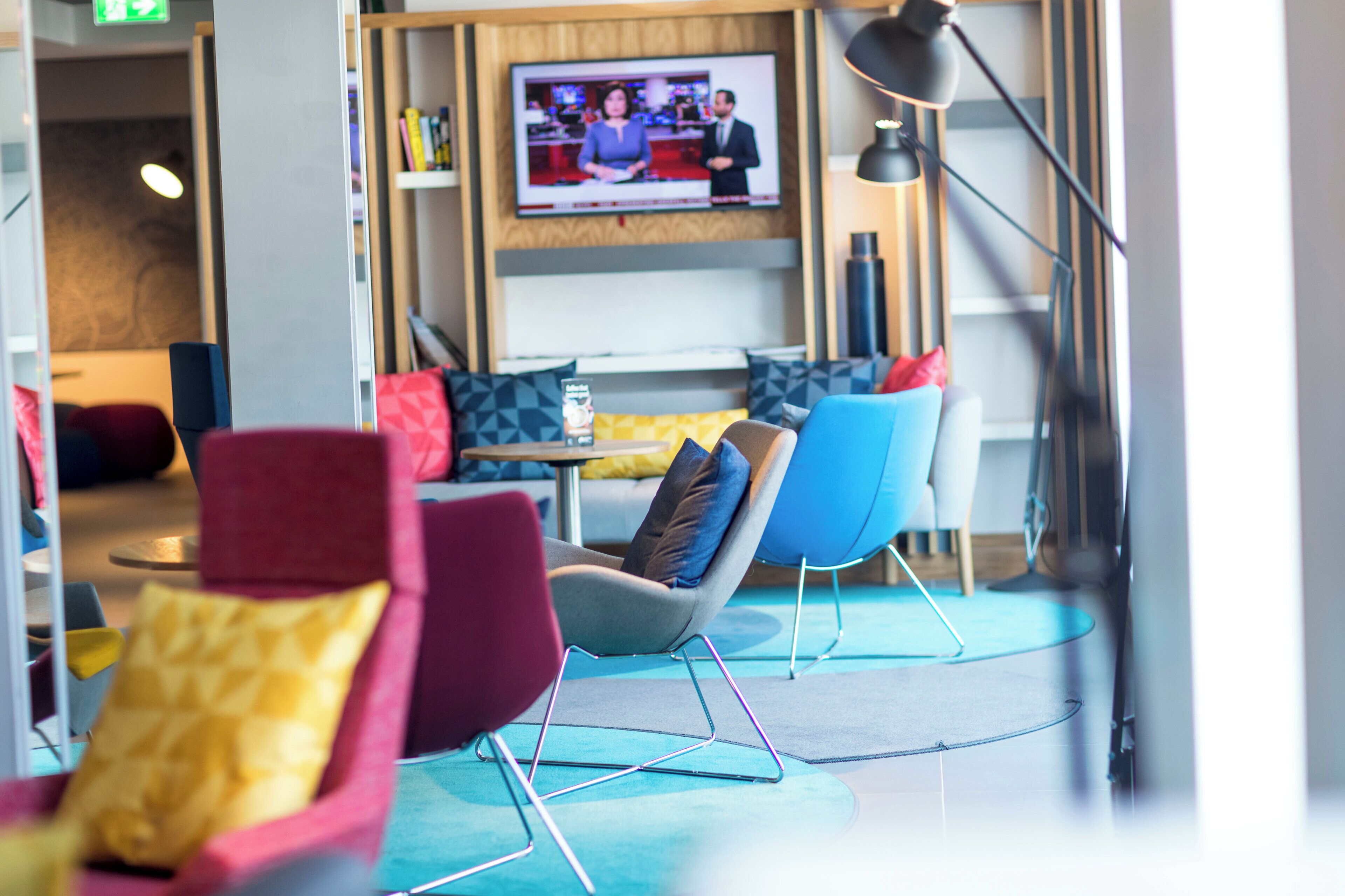 Eine moderne Lounge mit bunten Sitzmöglichkeiten und einem Fernseher, der eine Nachrichtensendung zeigt.