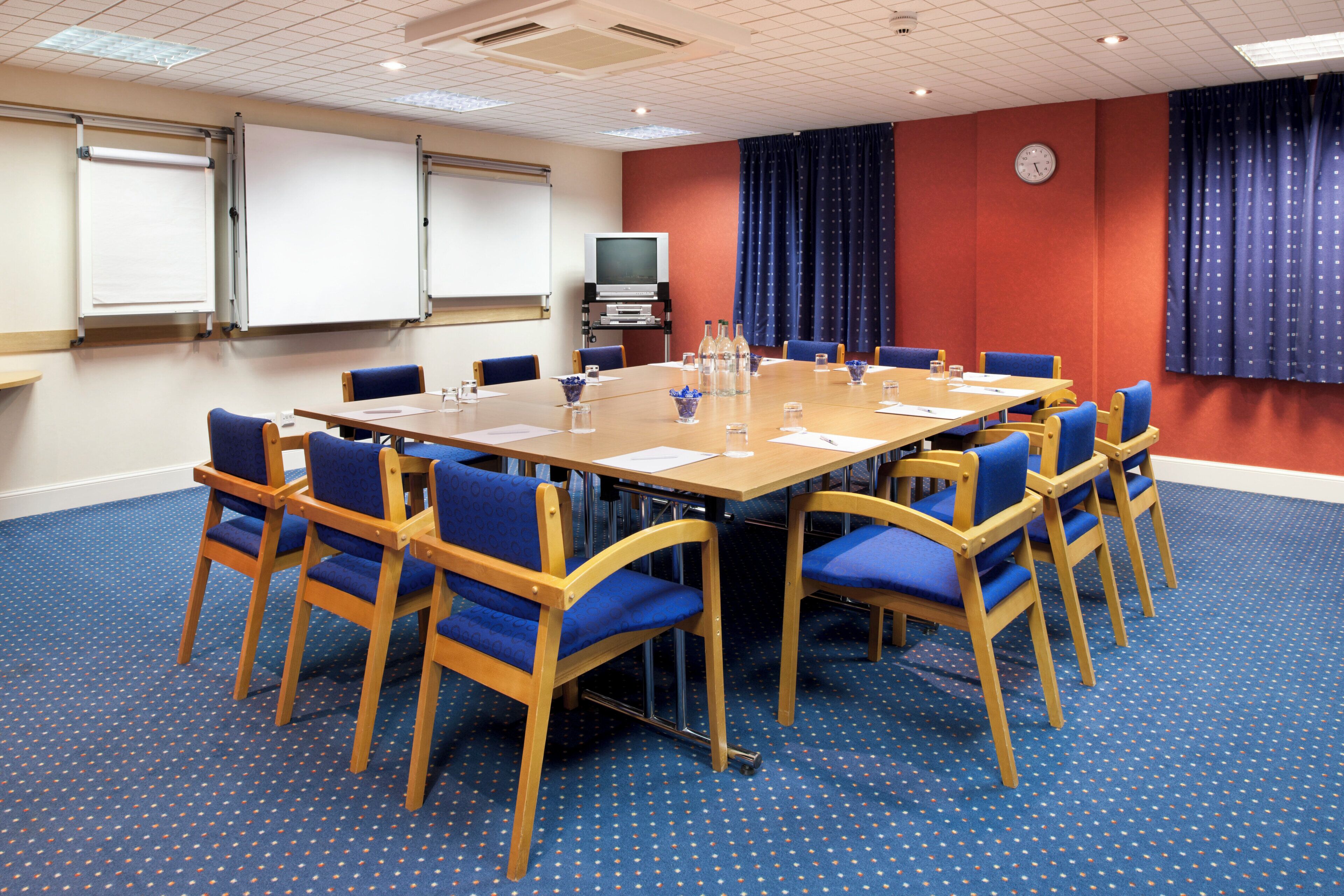 A conference room with a large table, blue chairs, a whiteboard, a clock, and a television on a stand.