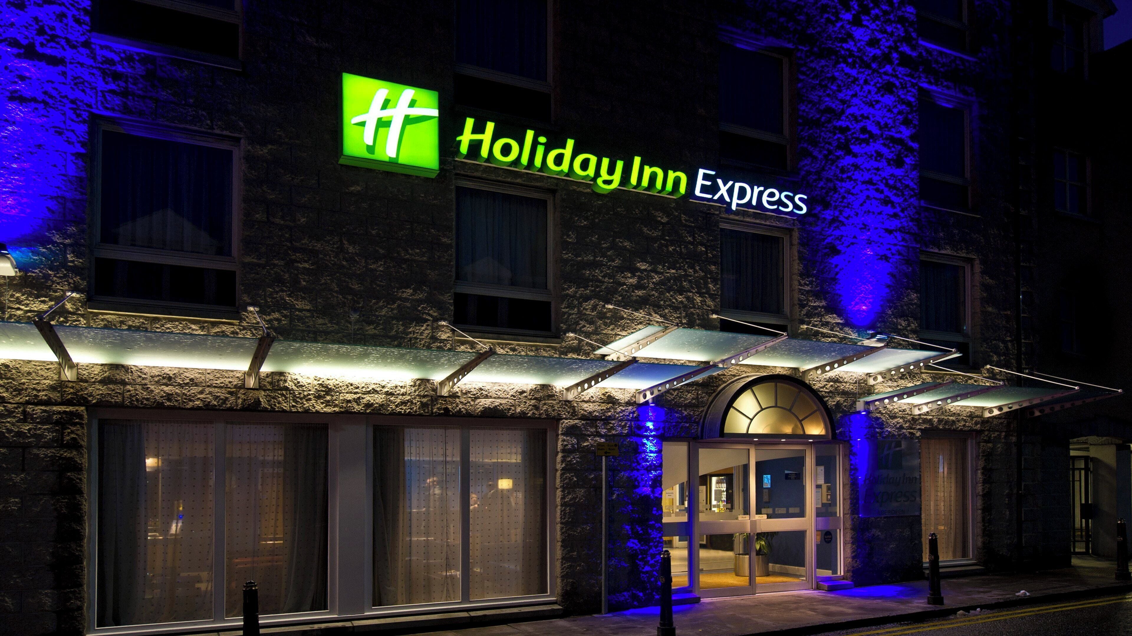 Edifício Holiday Inn Express com fachada de pedra, grandes janelas e entrada destacada com marquise.