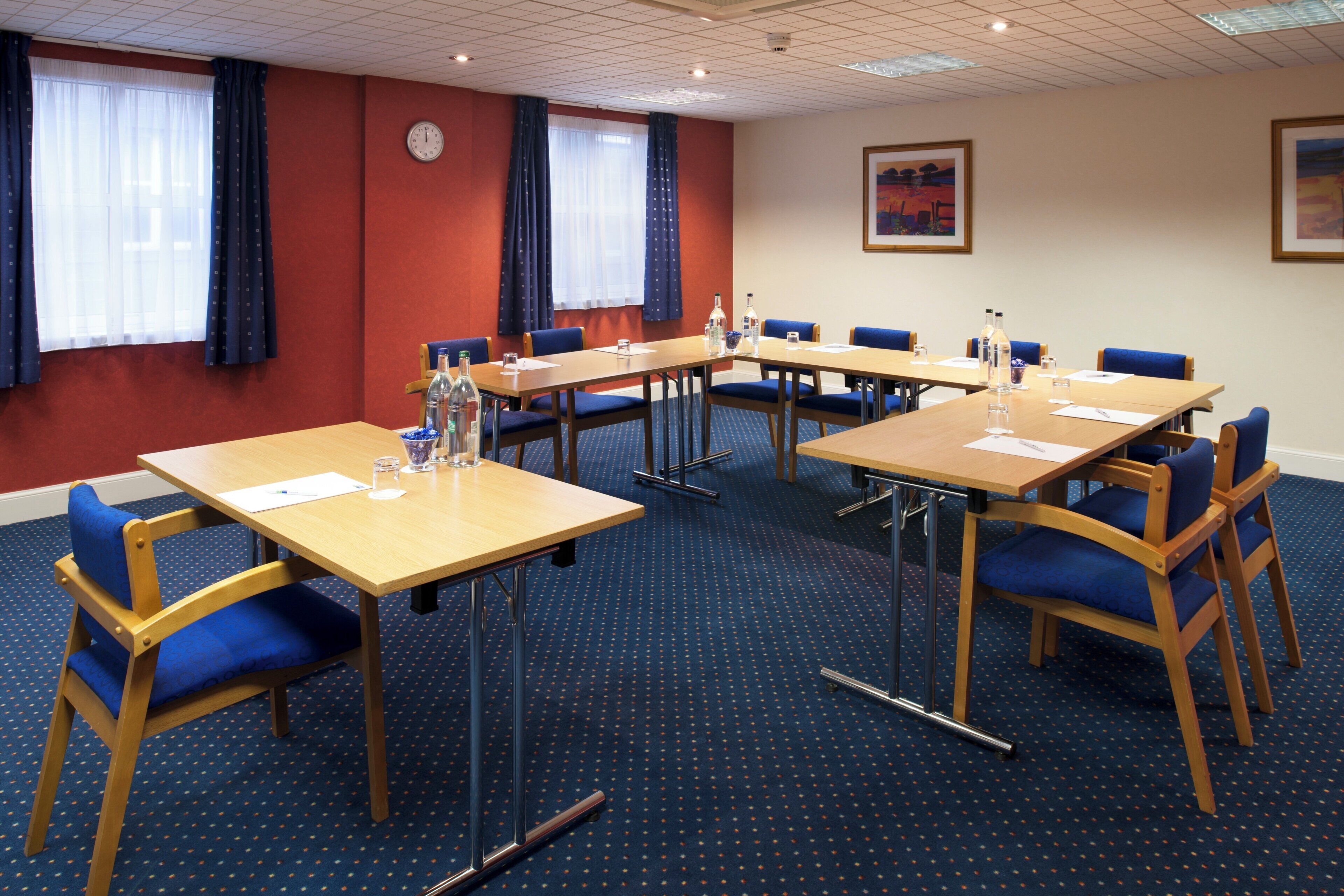 A conference room with multiple tables and chairs, a clock on the wall, and a painting above the door.