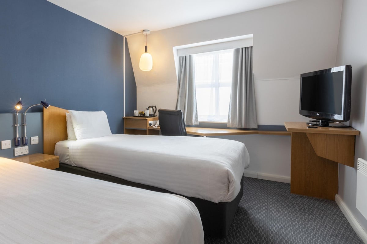Et hotelværelse med to senge, et skrivebord, et fjernsyn og et vindue med gardiner.