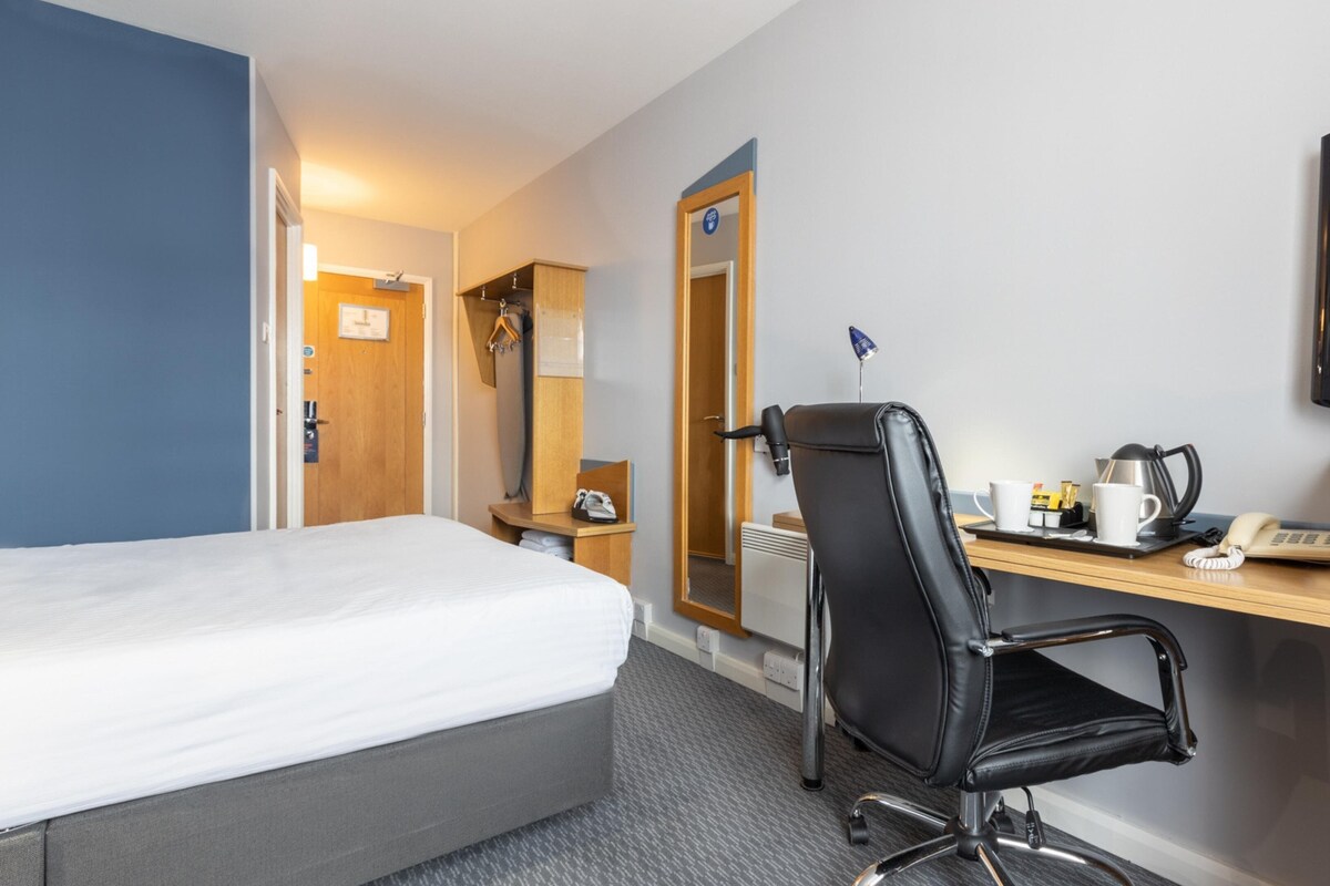Et hotelværelse med en seng, et skrivebord med stol, et fjernsyn og et spejl.