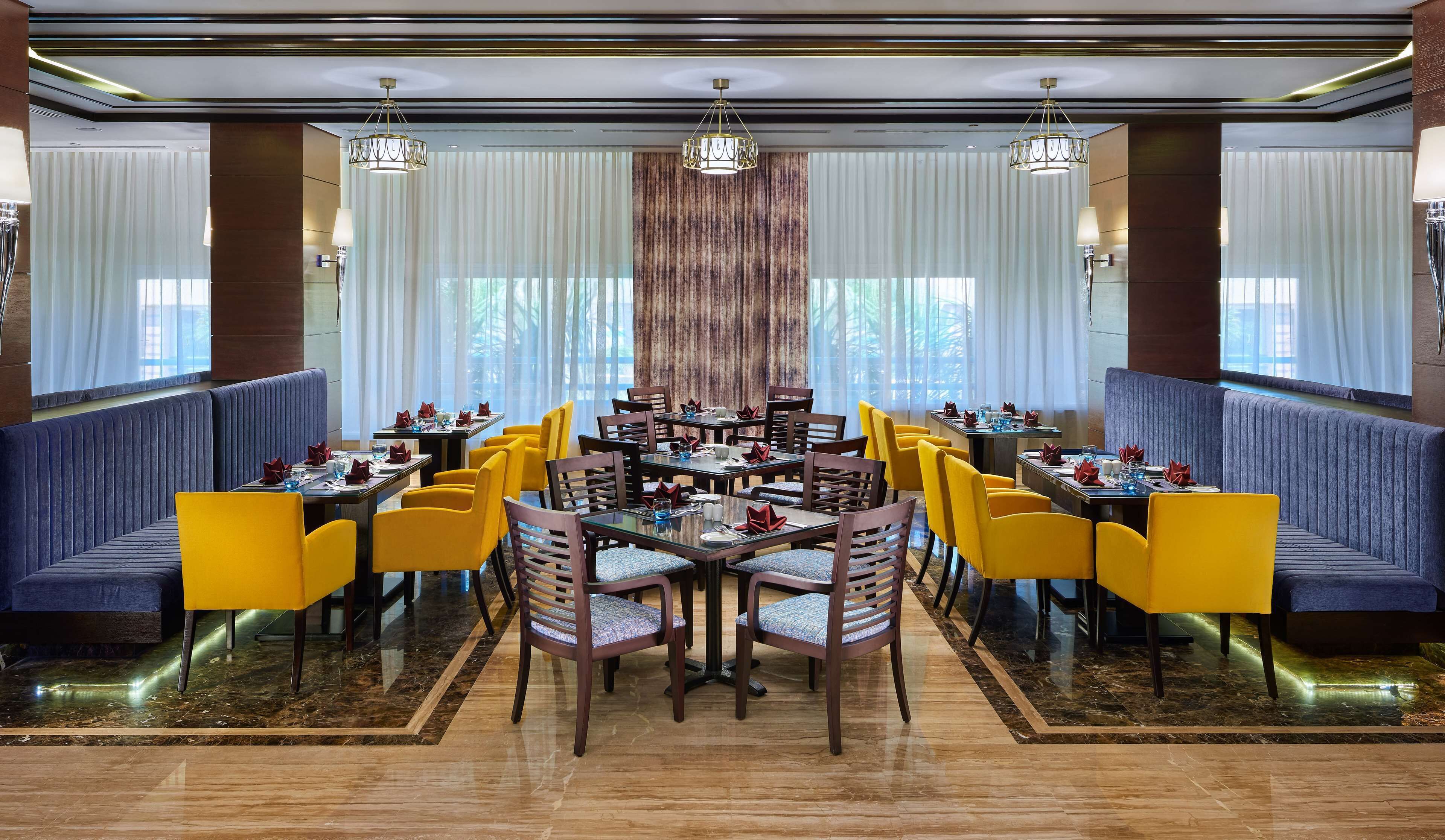 A hotel dining area with multiple tables and chairs, large windows with curtains, and decorative lighting.