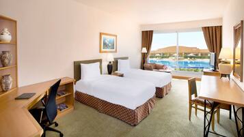 A hotel room with two beds, a desk, a chair, a TV, a bookshelf, and a large window with a pool view.