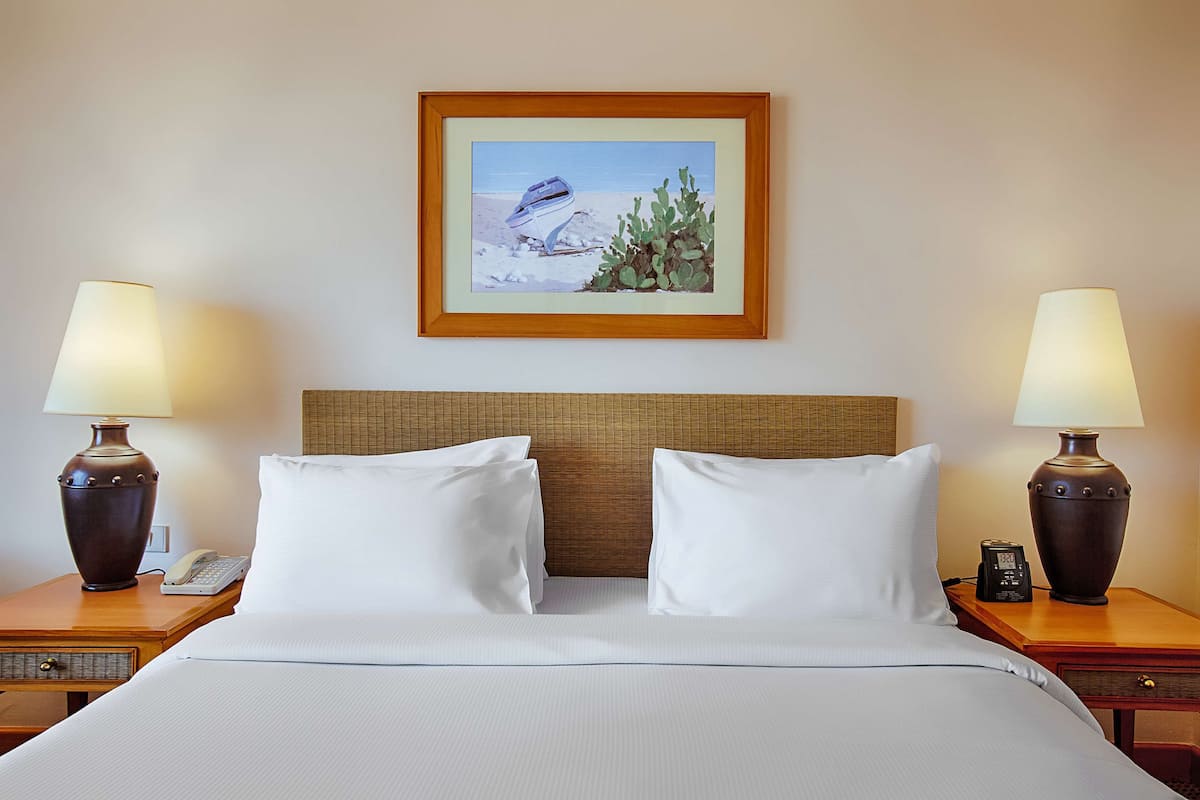 Una habitación de hotel con una cama, dos mesitas de noche con lámparas, un cuadro enmarcado en la pared y un buró con un teléfono.
