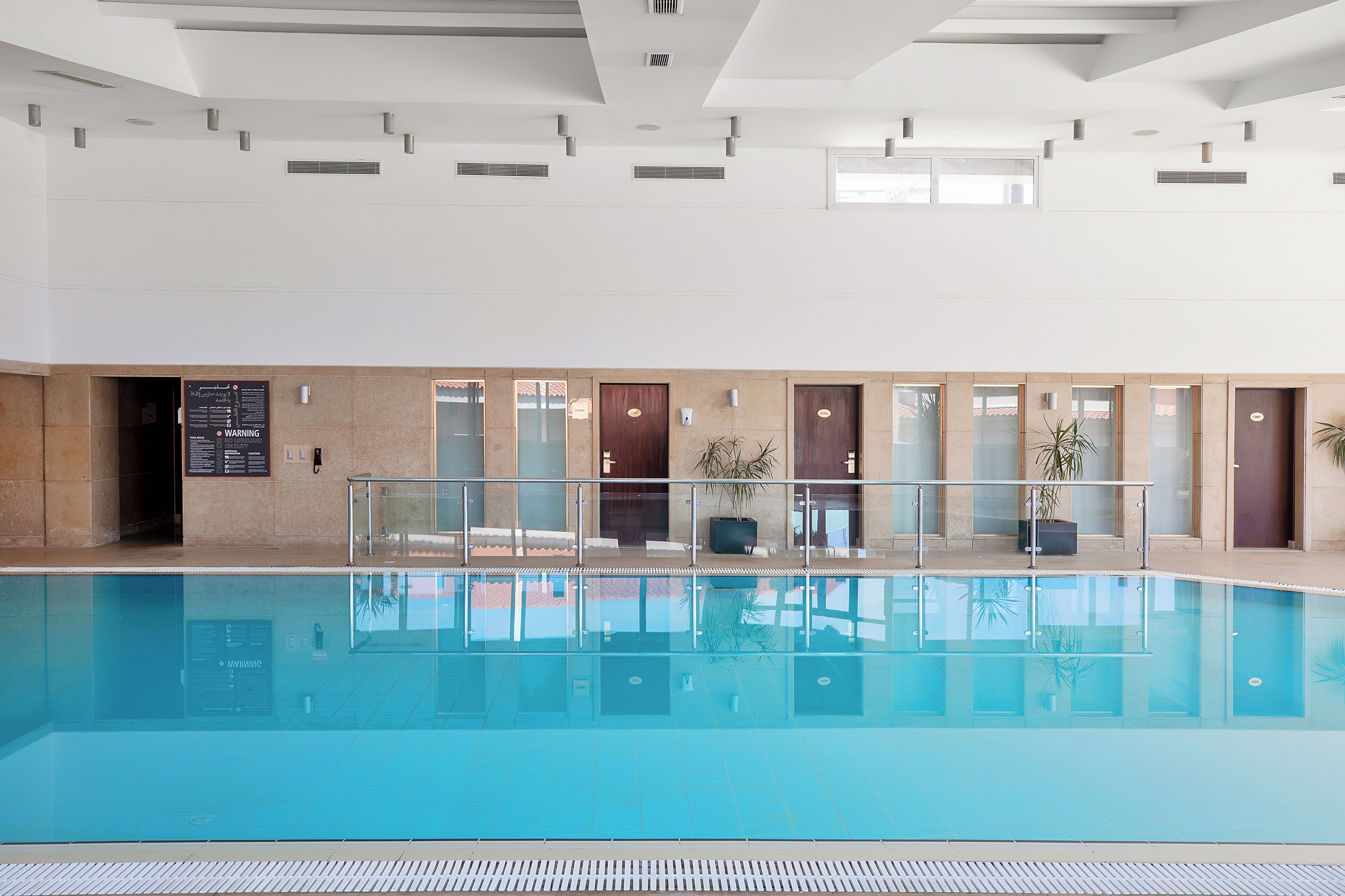 Une grande piscine intérieure à l’eau claire, entourée de murs et de portes de couleur beige, et dotée d’un sol carrelé.