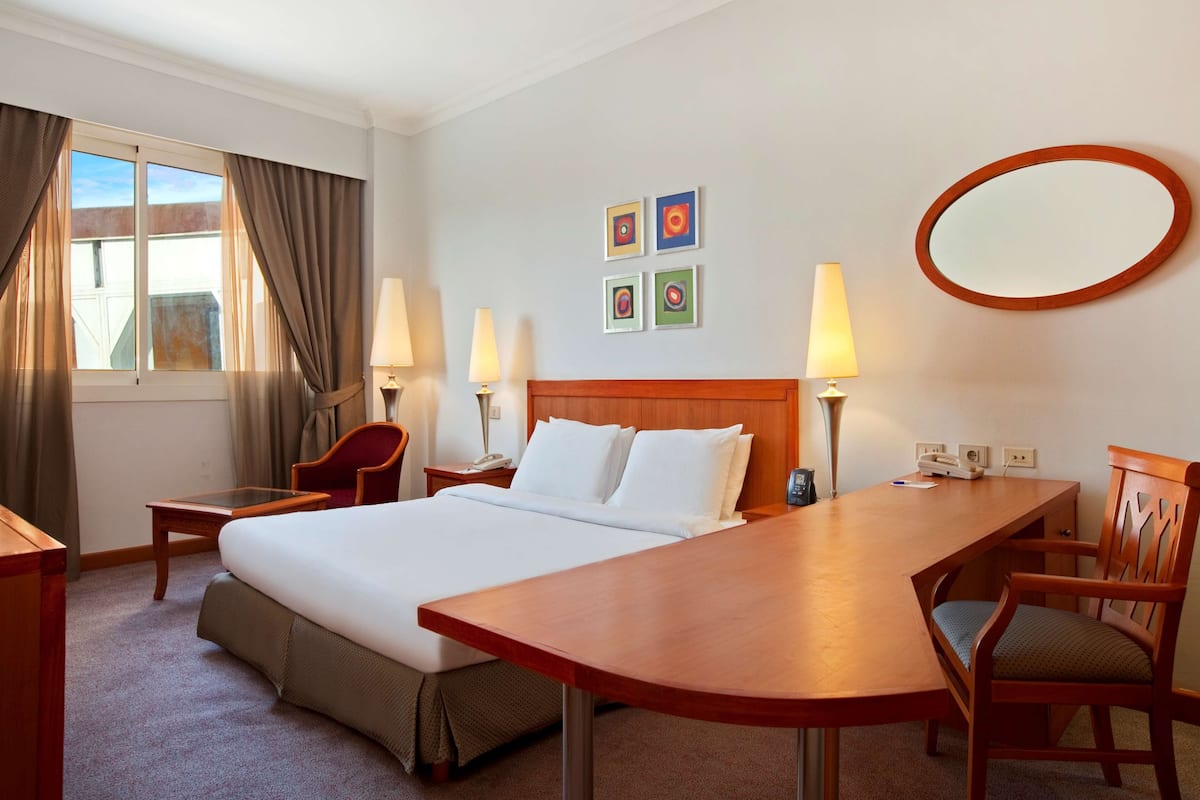 Una habitación de hotel con una cama grande, dos mesitas de noche con lámparas, un escritorio de madera, una silla y un espejo grande en la pared.
