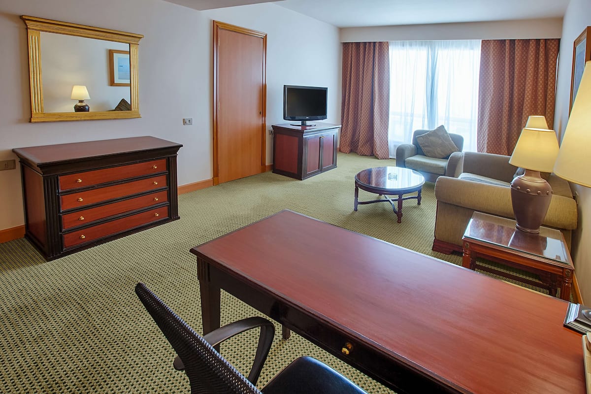 Habitación de hotel con cómoda de madera, televisor sobre un mueble bajo, sofá, mesa de centro y lámpara.