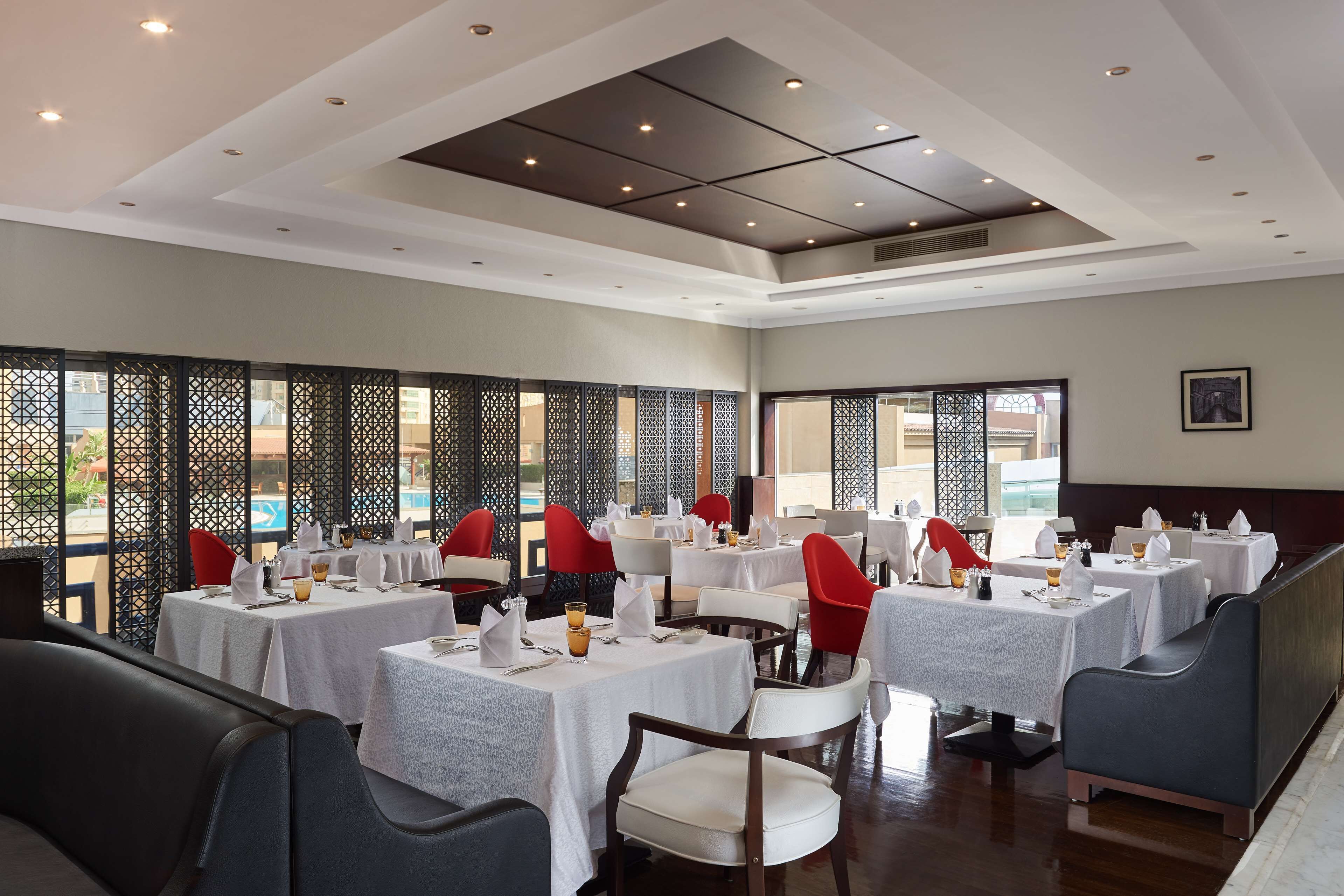 A restaurant with multiple tables and chairs, some with red cushions, and a ceiling with recessed lighting.