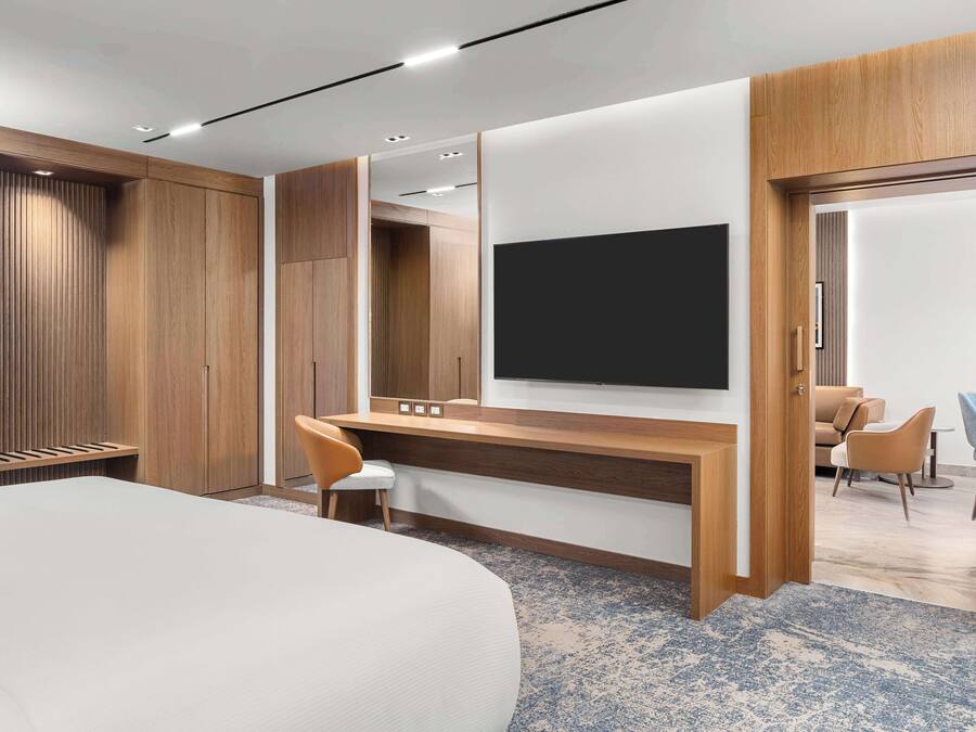 A modern hotel room with a large bed, a wooden desk with a flat-screen TV, and a seating area with chairs.