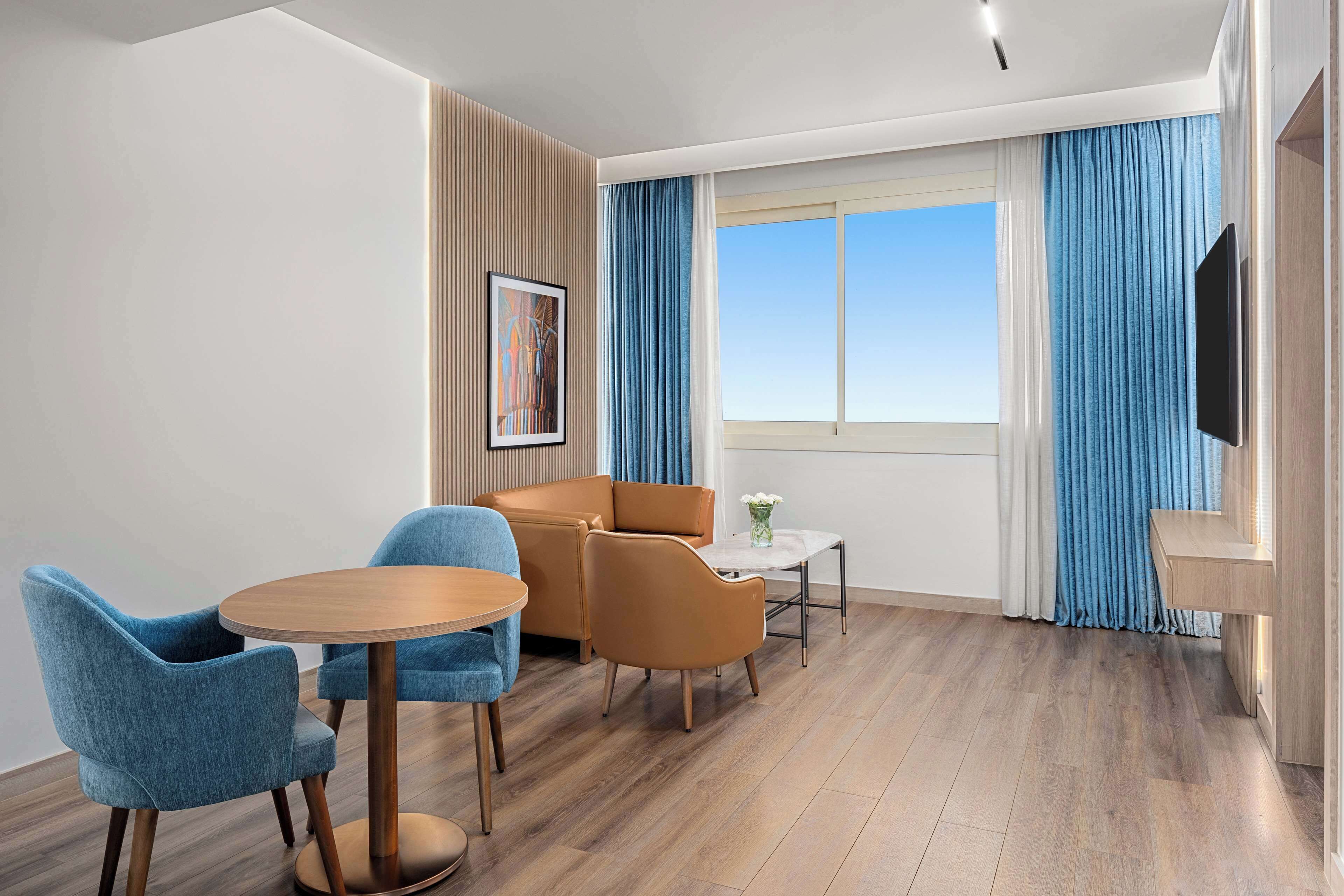 Una habitación de hotel moderna con suelo de madera, una mesa redonda con sillas, un sofá, un televisor de pantalla plana y un ventanal con cortinas.