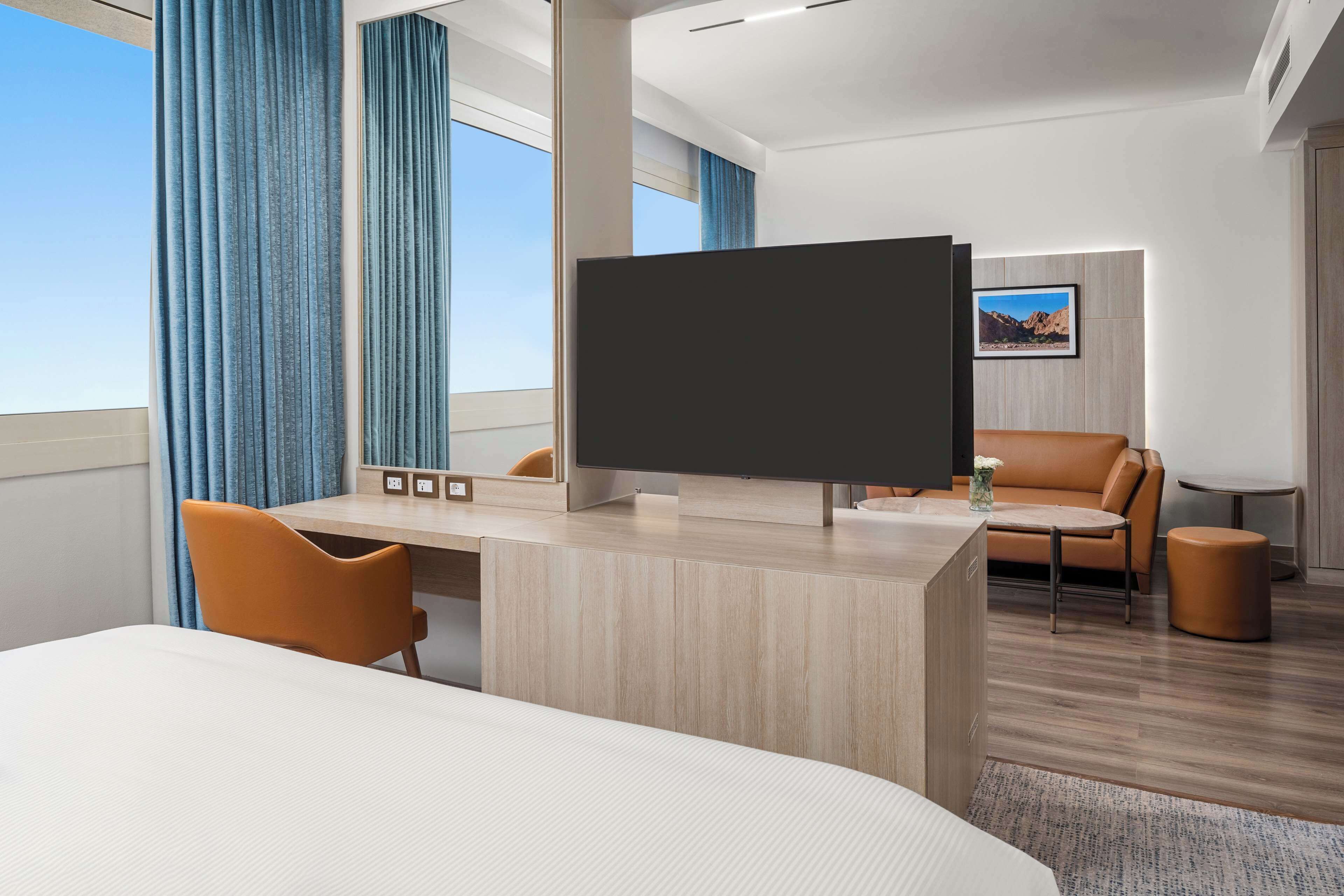 A modern hotel room with a large bed, a desk with a flat-screen TV, a leather sofa, and a small round table.
