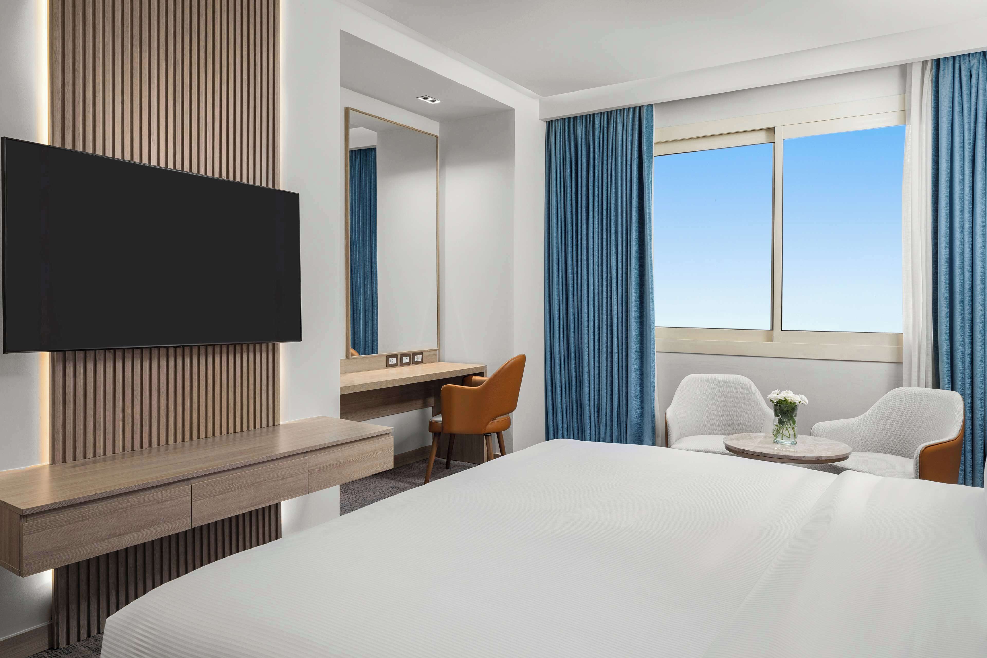 Habitación de hotel con una cama grande, un escritorio con silla, un televisor montado en la pared y una ventana con cortinas.