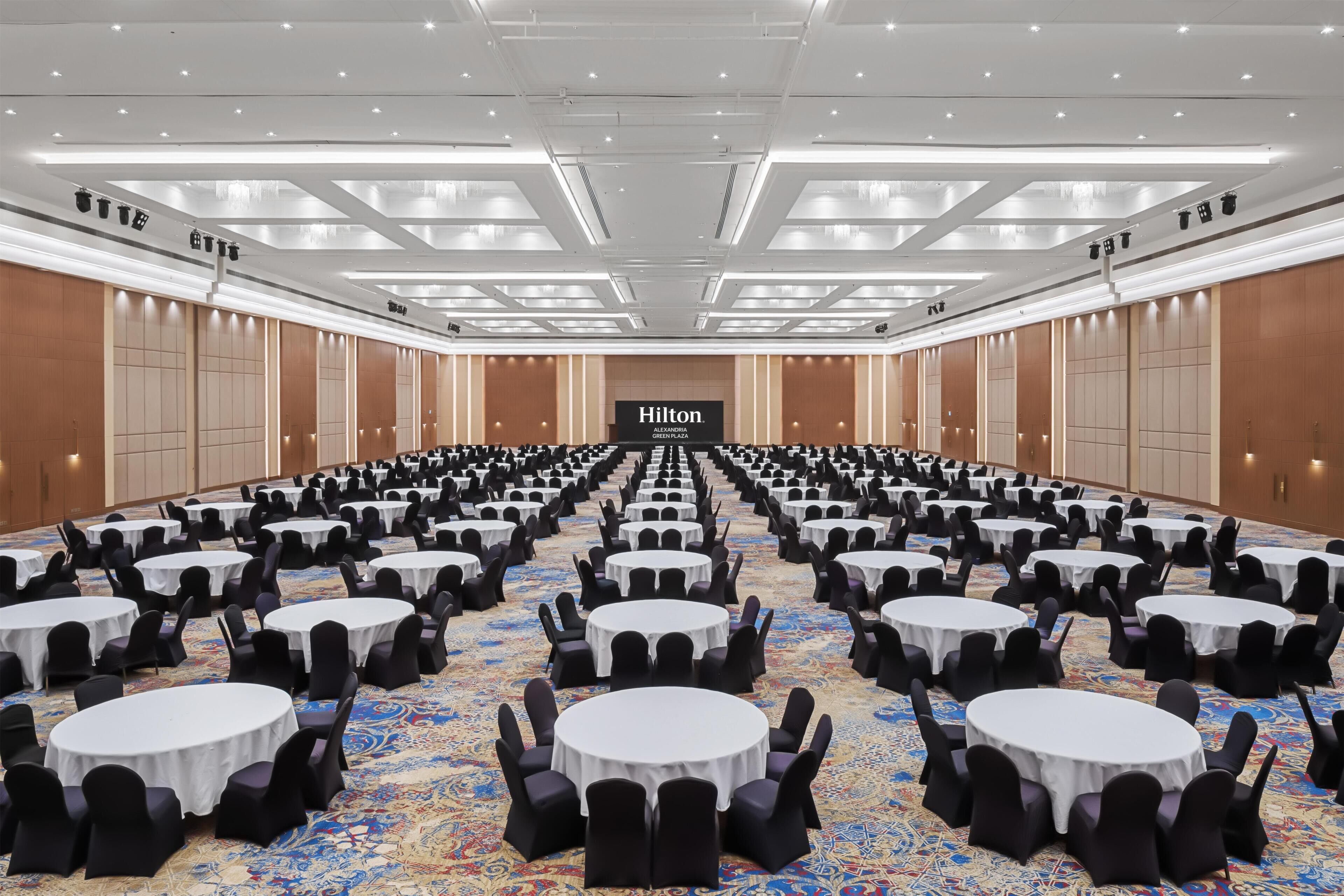 A large, well-lit conference room with rows of round tables and chairs, a visible Hilton sign, and a patterned carpet.