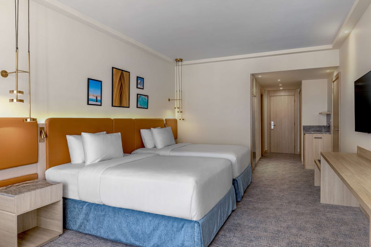 Habitación de hotel con dos camas, un escritorio, un televisor y una puerta que da a un pasillo.