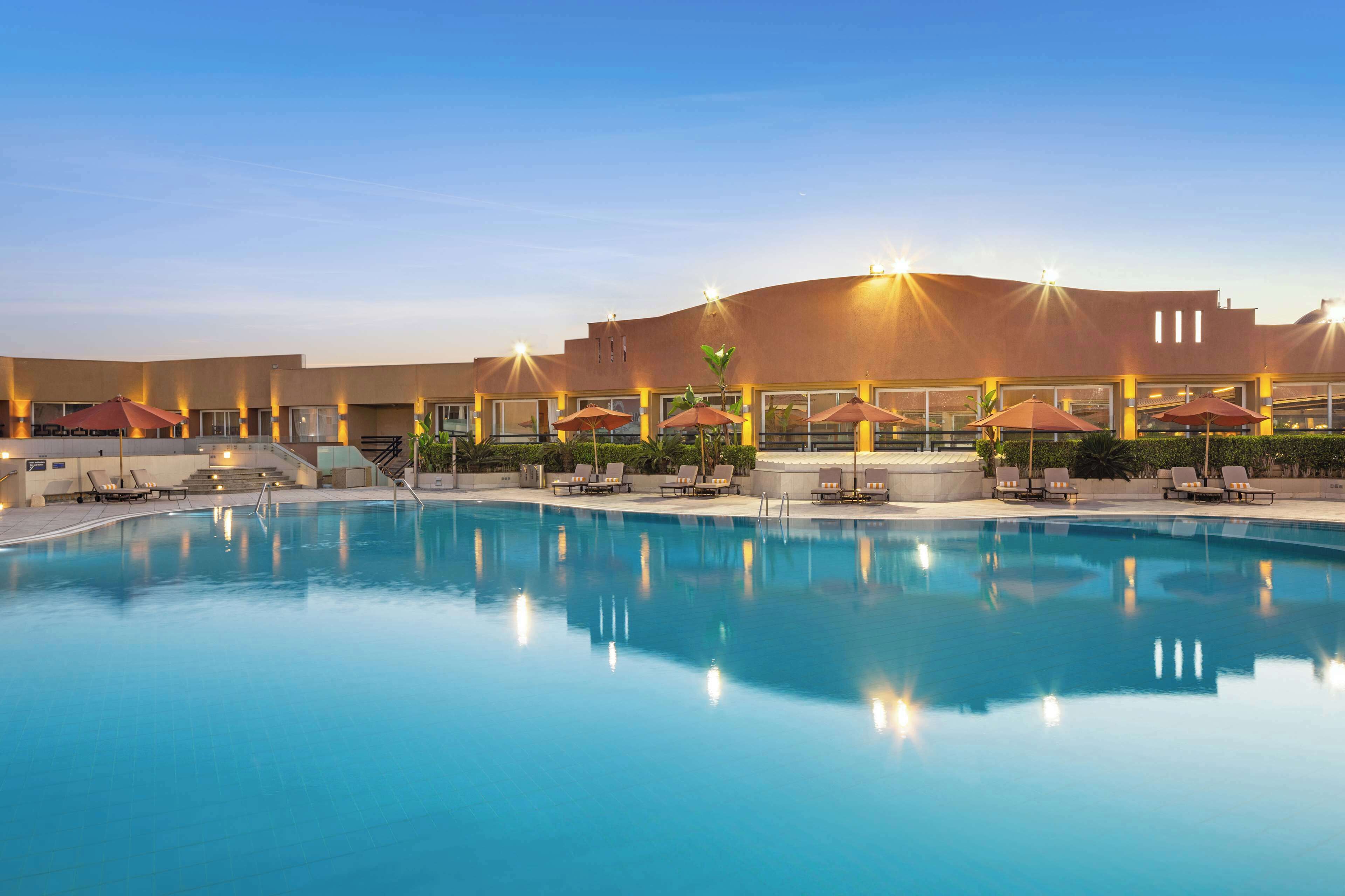 Une piscine entourée de chaises longues et de parasols, le tout bordé par un bâtiment doté de nombreuses fenêtres et d’un toit plat.