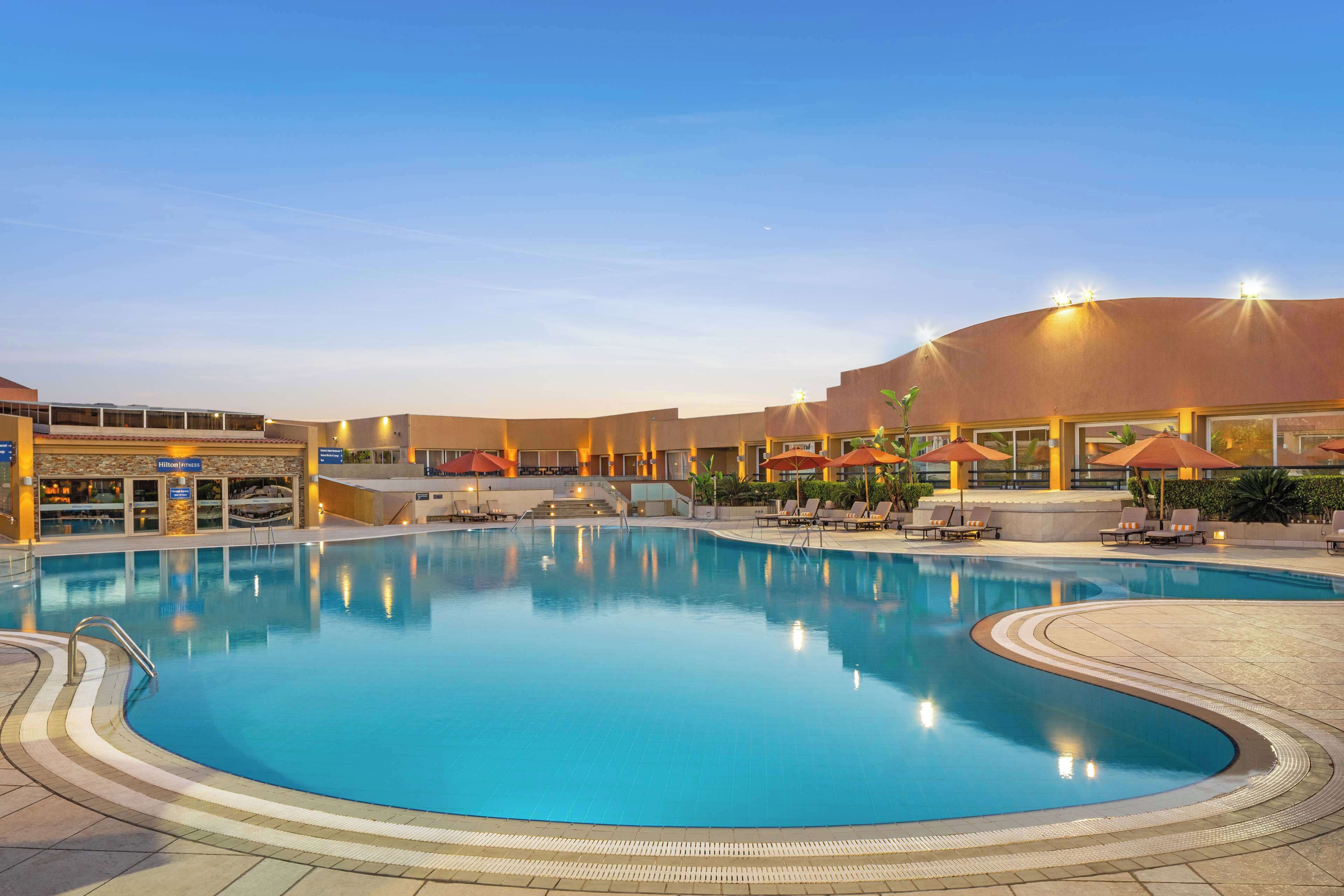 A large indoor swimming pool with a curved edge, surrounded by lounge chairs and umbrellas, and a building with a sign that reads "Hilton".