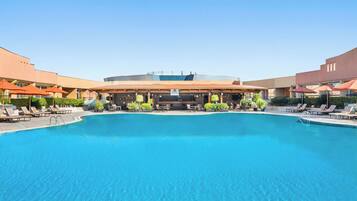 A large outdoor swimming pool with lounge chairs and umbrellas, surrounded by a building with a flat roof and a covered seating area.