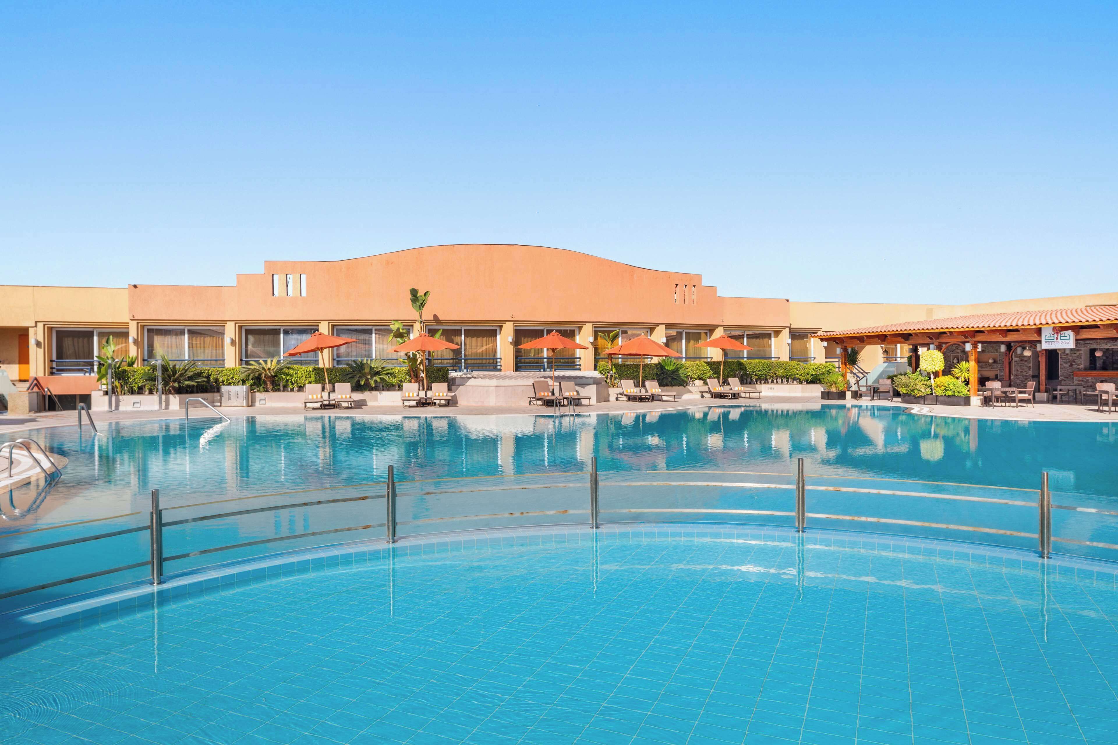 A large swimming pool with sun loungers and umbrellas, surrounded by a building with a curved roof and multiple windows.