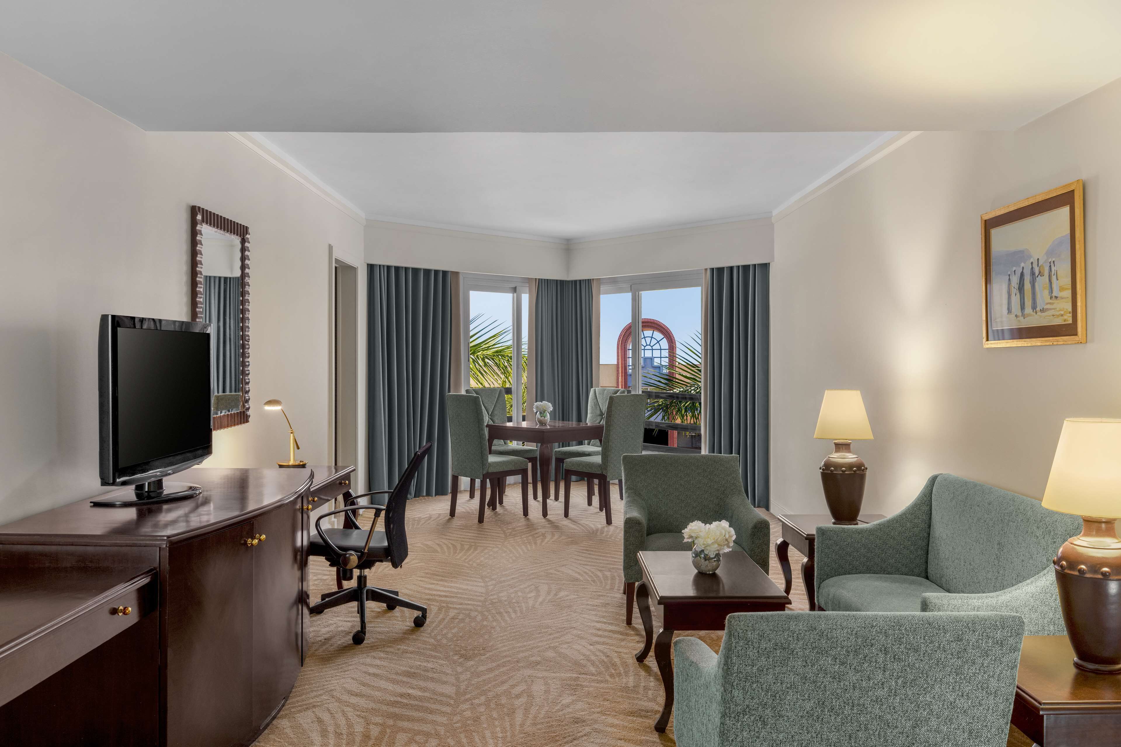 A hotel room with a TV, desk, two armchairs, a dining table with chairs, and a view of a building through large windows.