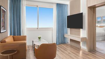 Habitación de hotel moderna con sofá, una mesa de comedor con sillas, televisor montado en la pared y un ventanal con cortinas.