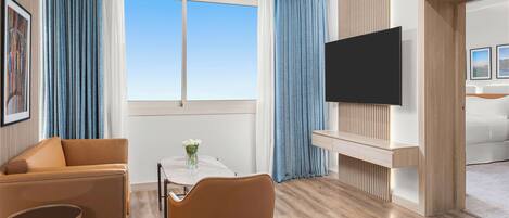 Habitación de hotel moderna con sofá, una mesa de comedor con sillas, televisor montado en la pared y un ventanal con cortinas.