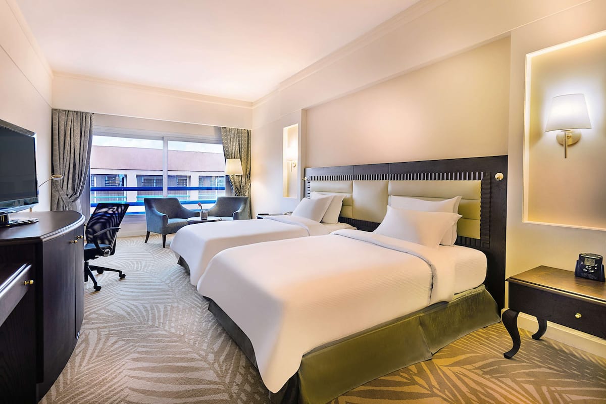 Habitación de hotel con dos camas, un escritorio con televisión, una silla y vistas a edificios a través de amplias ventanas.