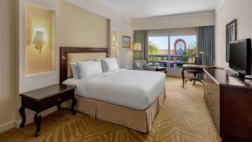 A hotel room with a large bed, a desk, a chair, a television, and a balcony with a view of palm trees and a building.