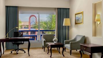 A hotel room with a desk, two armchairs, a small table with a vase, a lamp, and a painting on the wall.