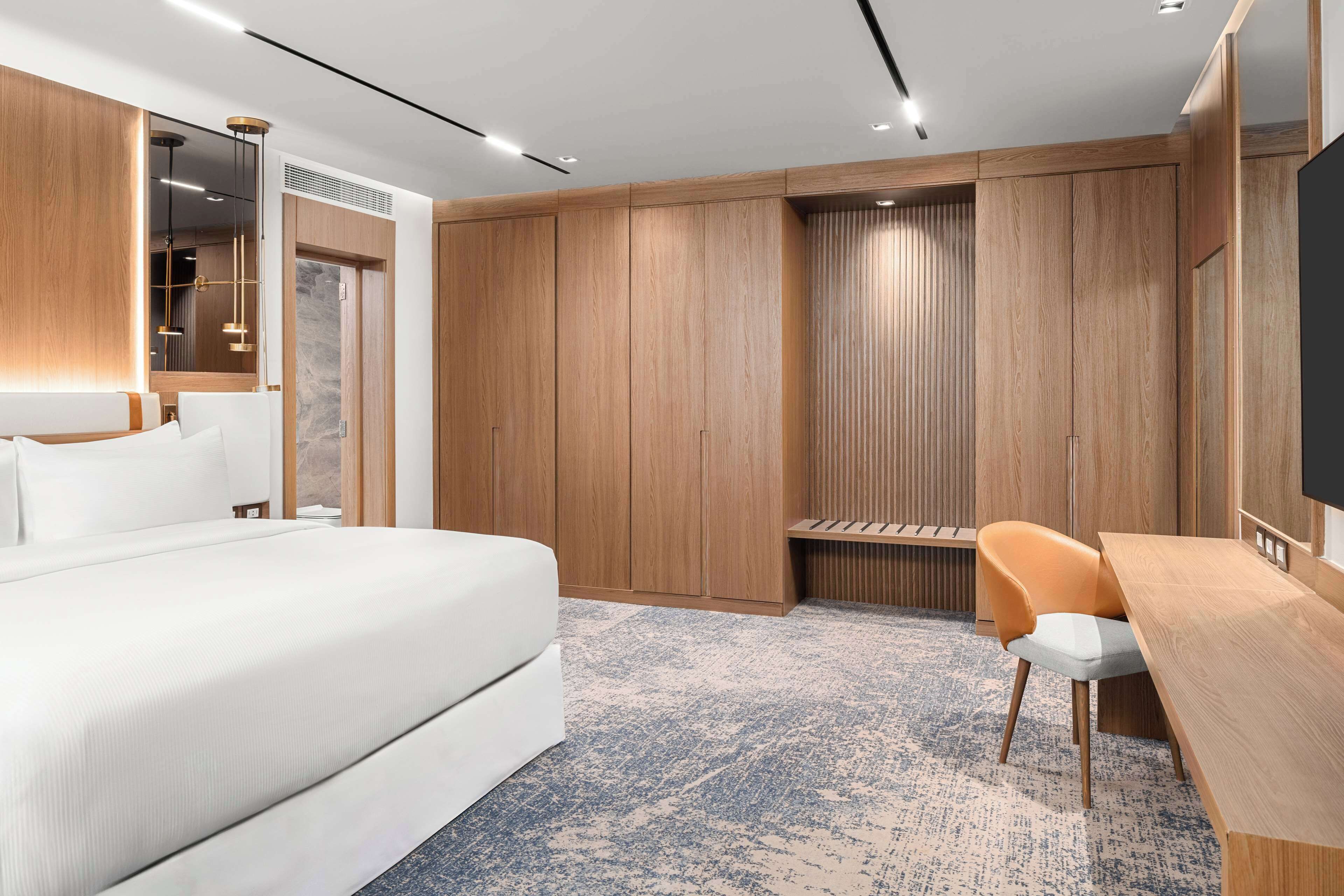 A modern hotel room with a large bed, a wooden wardrobe, a desk, and a chair.