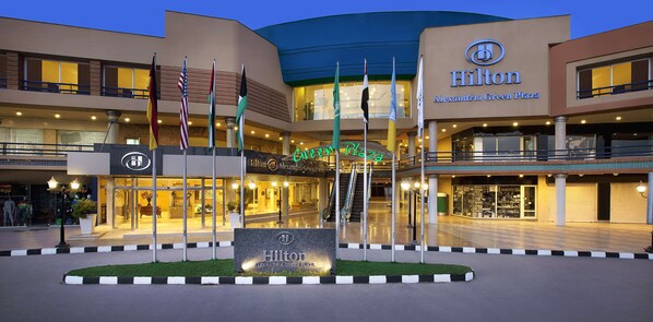 The exterior of the Hilton Alexandria Green Plaza hotel with multiple flags, a well-lit entrance, and a clear view of the surrounding area.