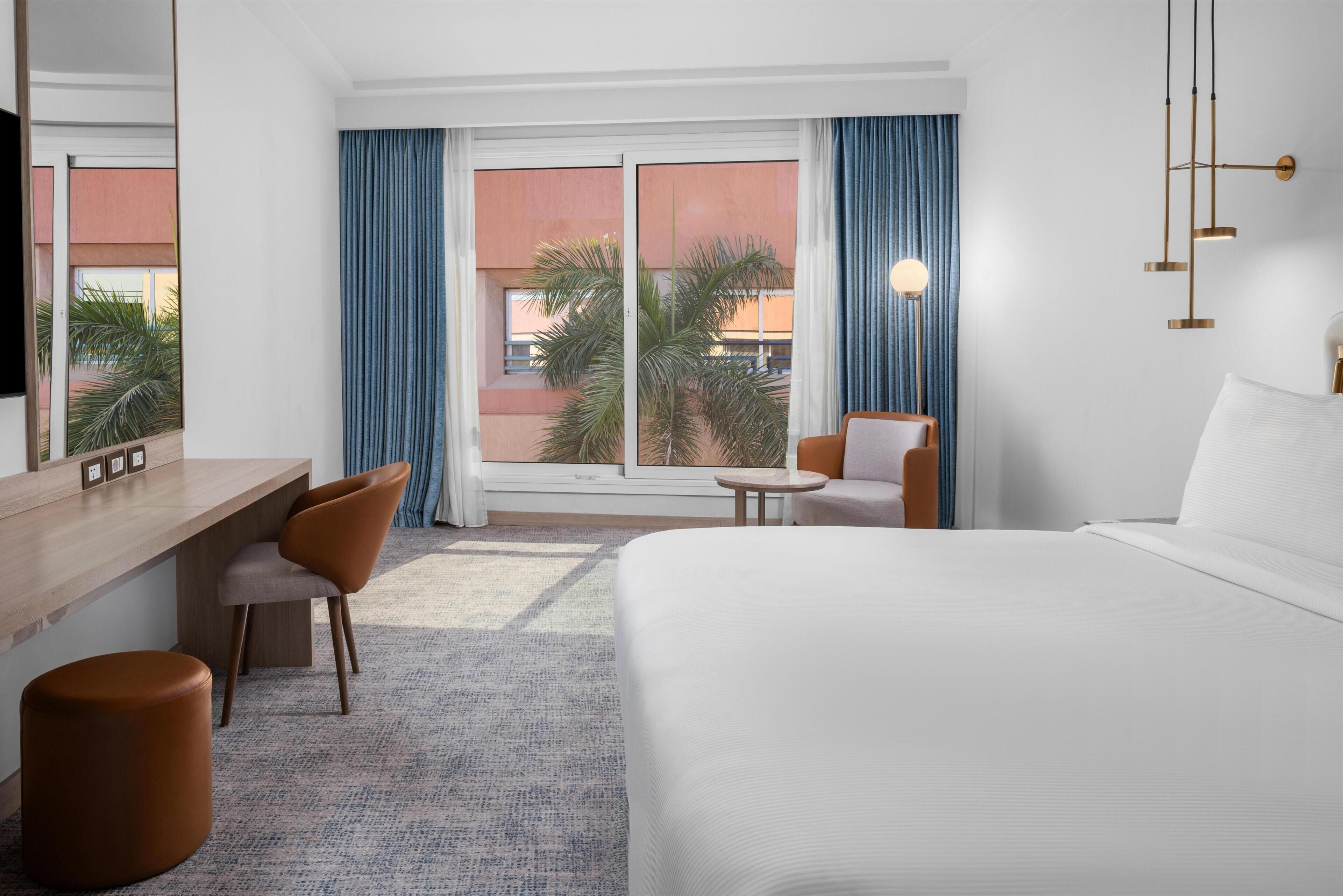 A hotel room with a bed, a desk, a chair, a round ottoman, and a large window with curtains.