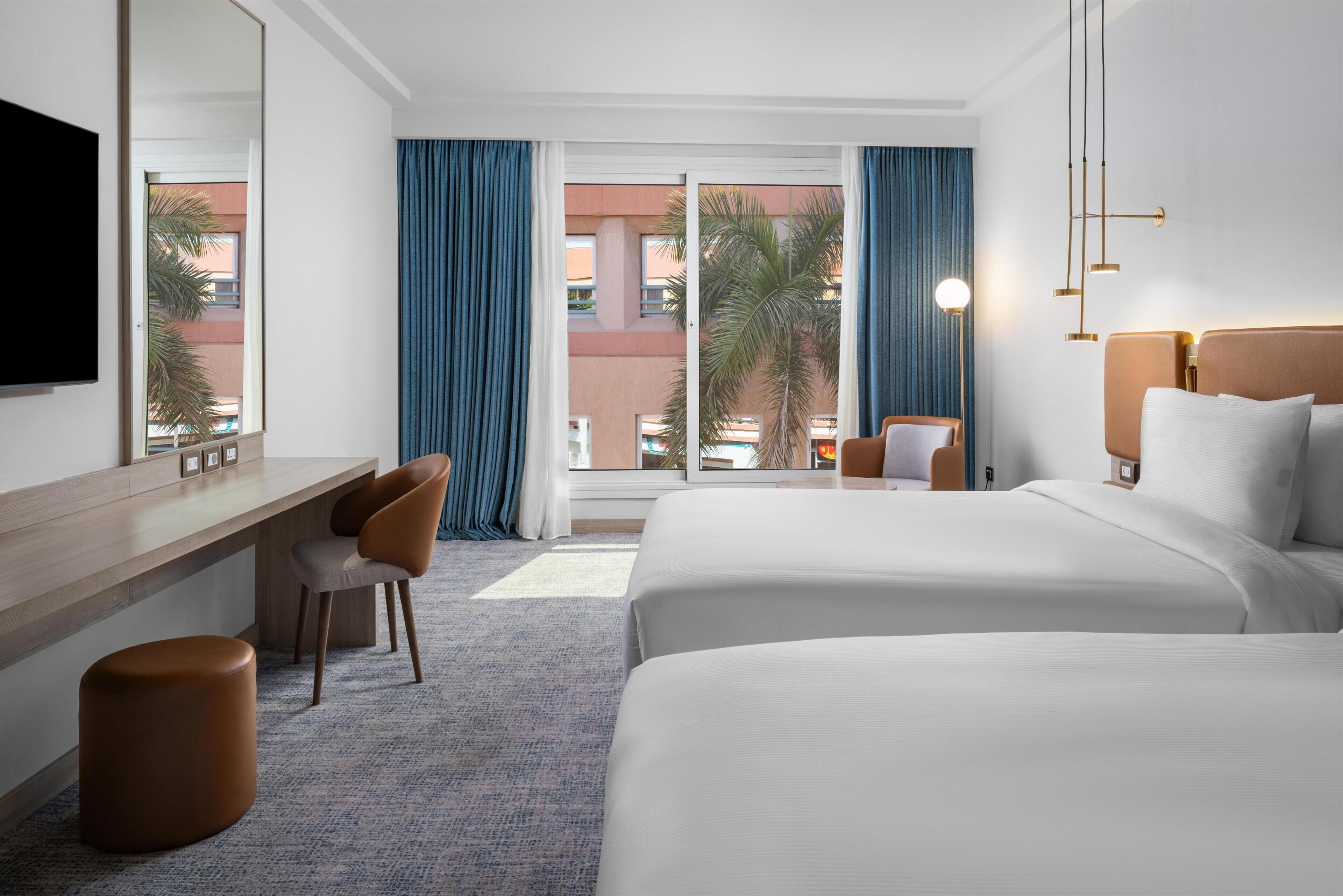 A hotel room with a large bed, a desk, a chair, a mirror, and a view of the outside.