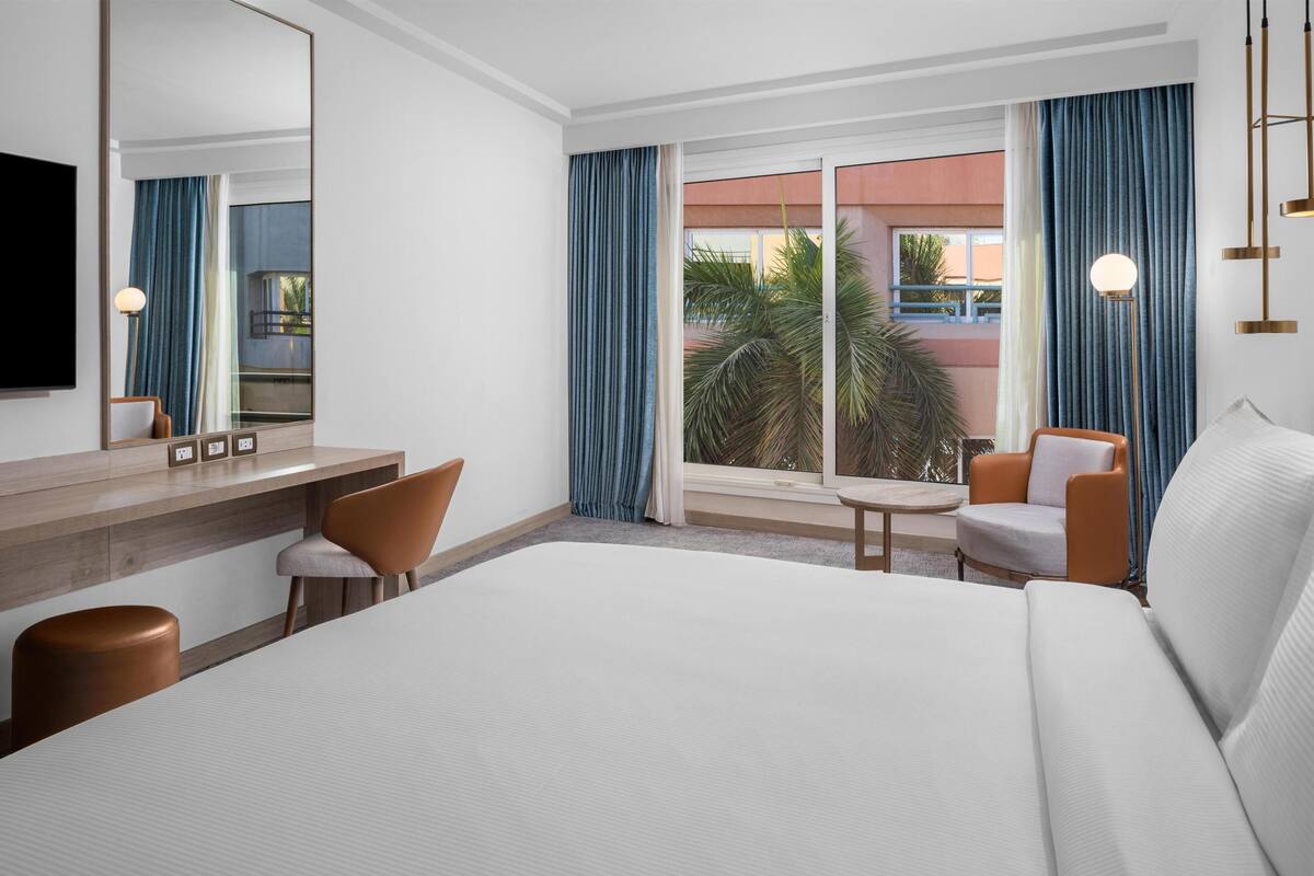 Habitación de hotel con una cama grande, un escritorio con una silla, una mesita pequeña, un sofá, un espejo amplio y vistas al exterior.