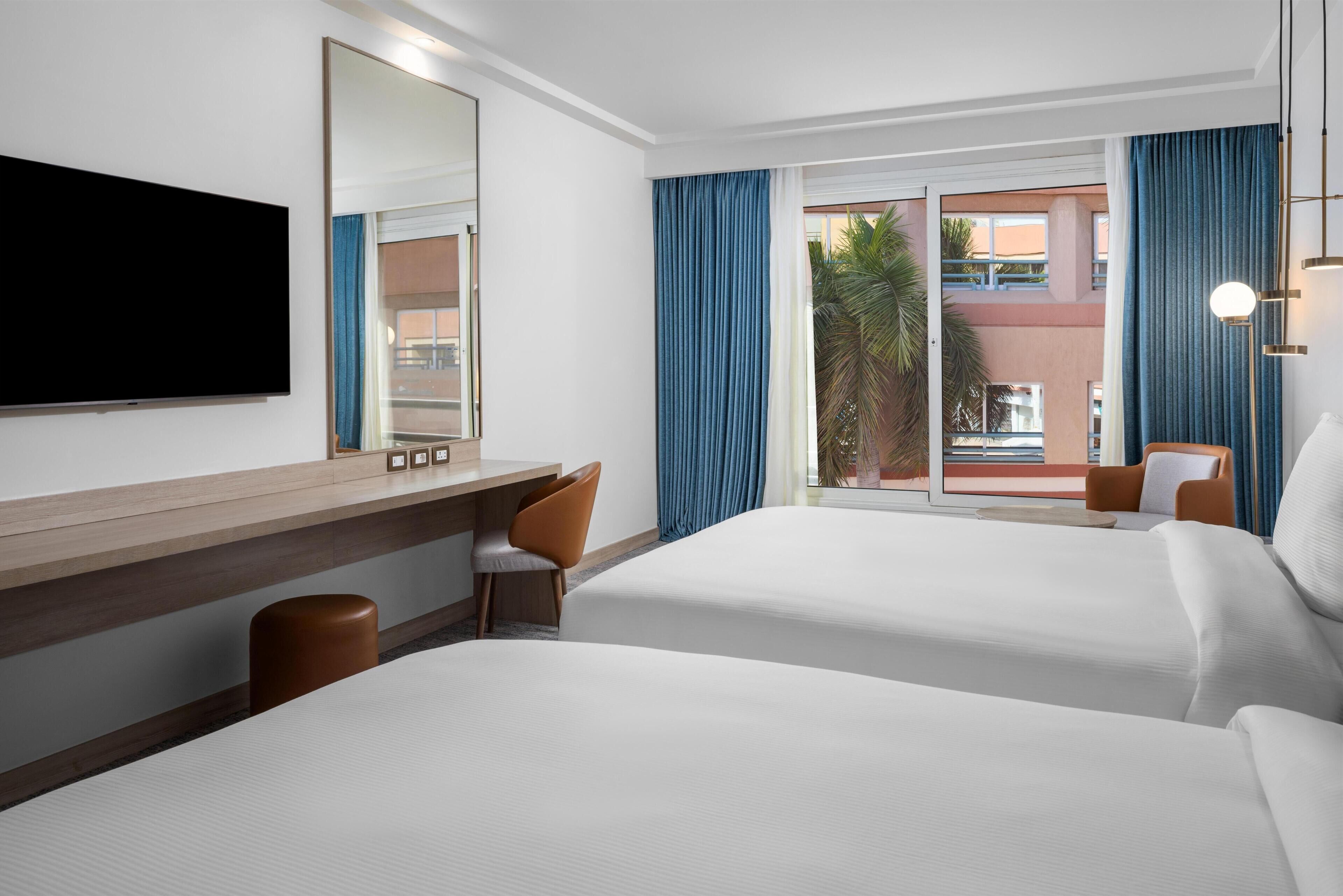 Habitación de hotel con dos camas, un escritorio, una silla, un televisor y un ventanal con cortinas.