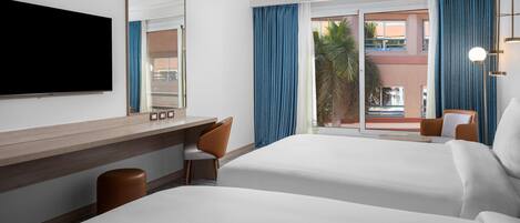 Habitación de hotel con dos camas, un escritorio, una silla, un televisor y un ventanal con cortinas.