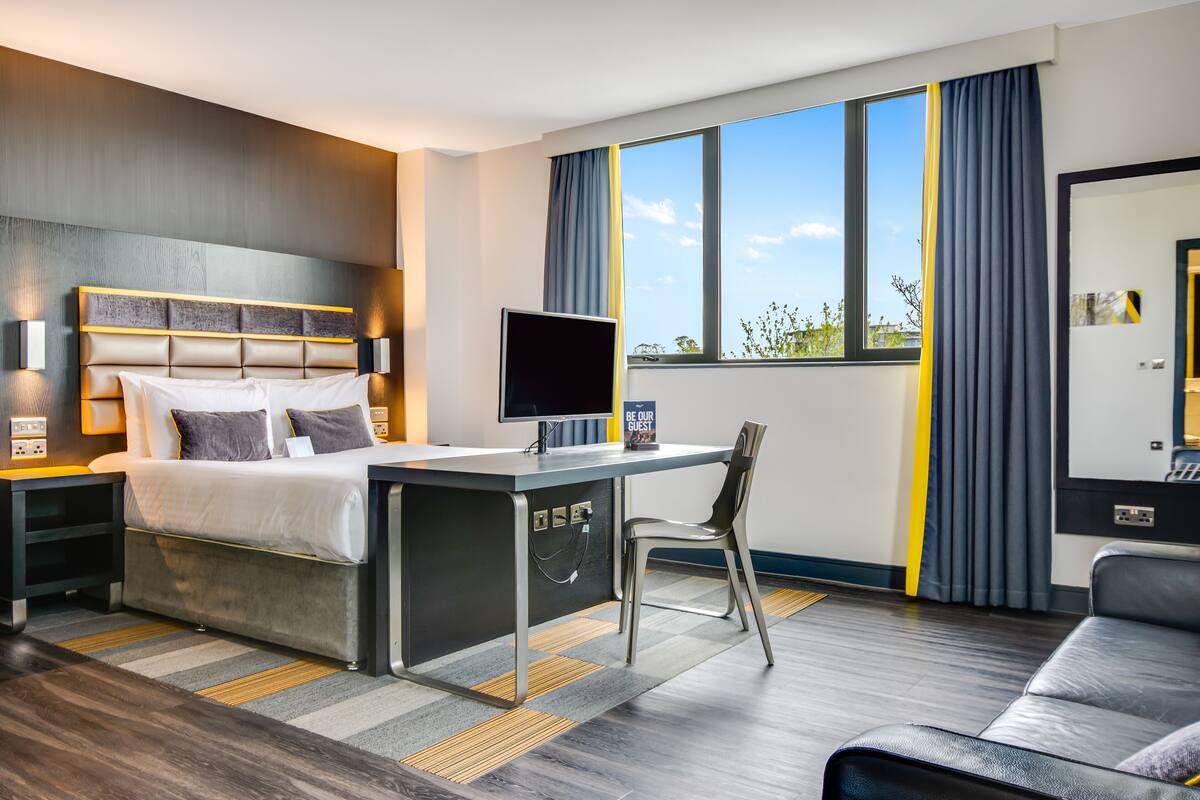 A modern hotel room with a bed, desk, and chair, large windows, and a view of the outdoors.
