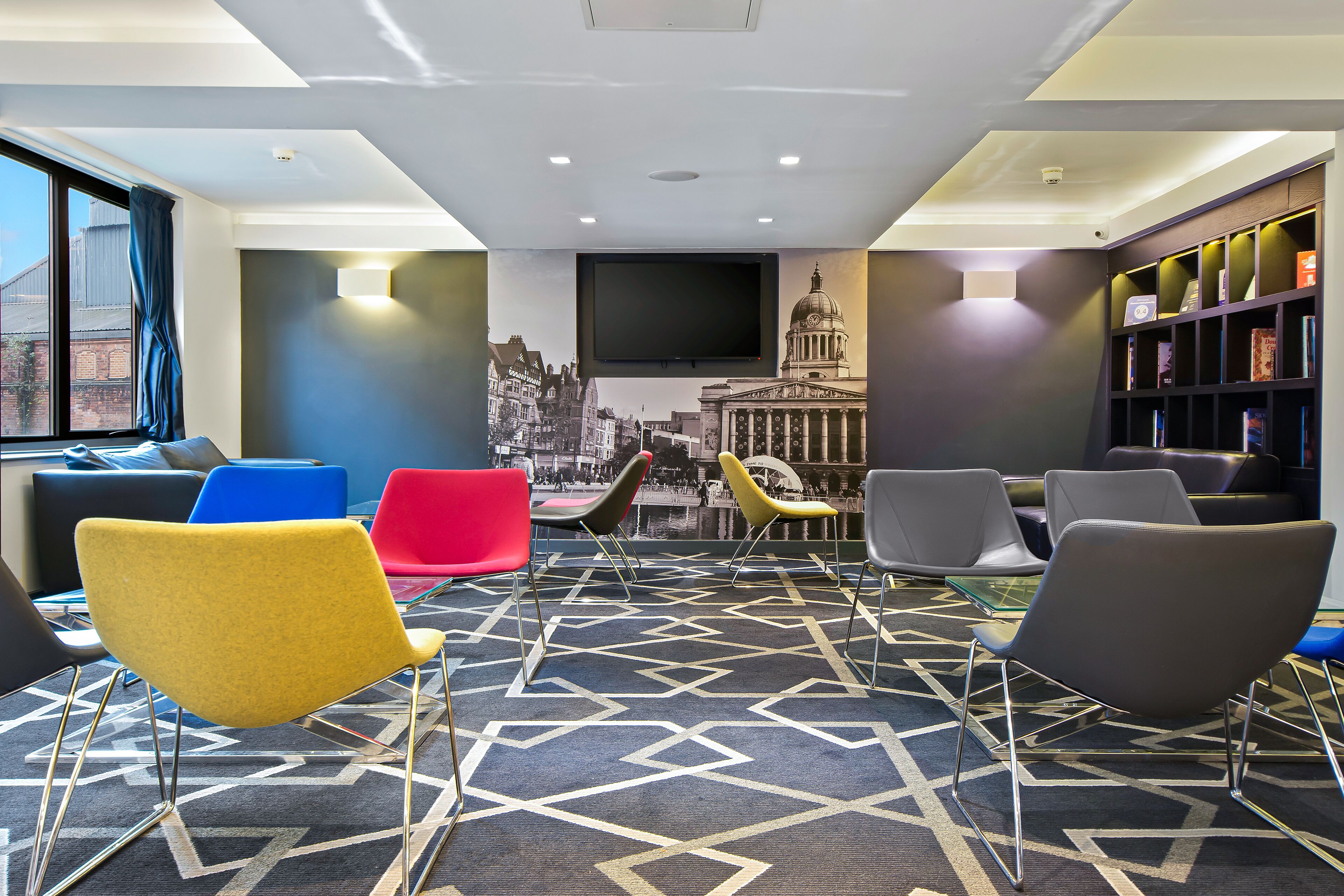 A modern waiting area with a large TV screen, colorful chairs, a patterned carpet, and a bookshelf.