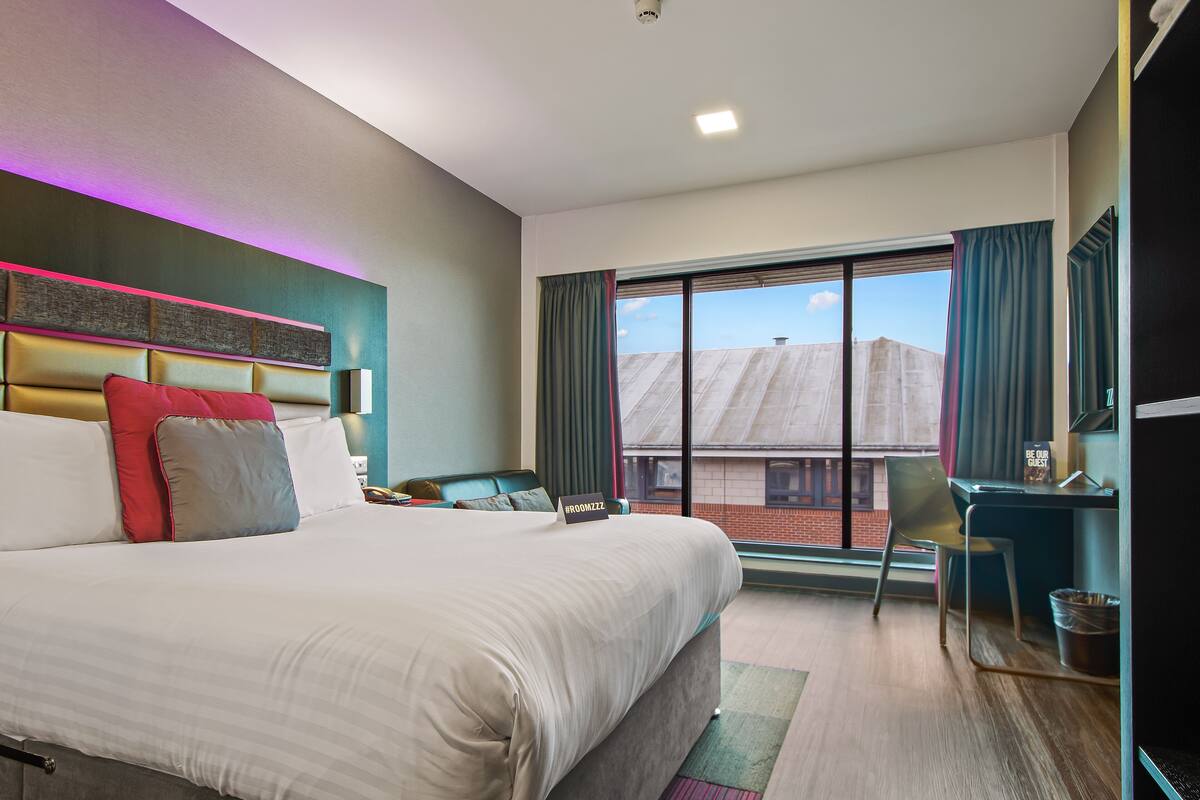A modern hotel room with a large bed, a desk, and a view of the cityscape.