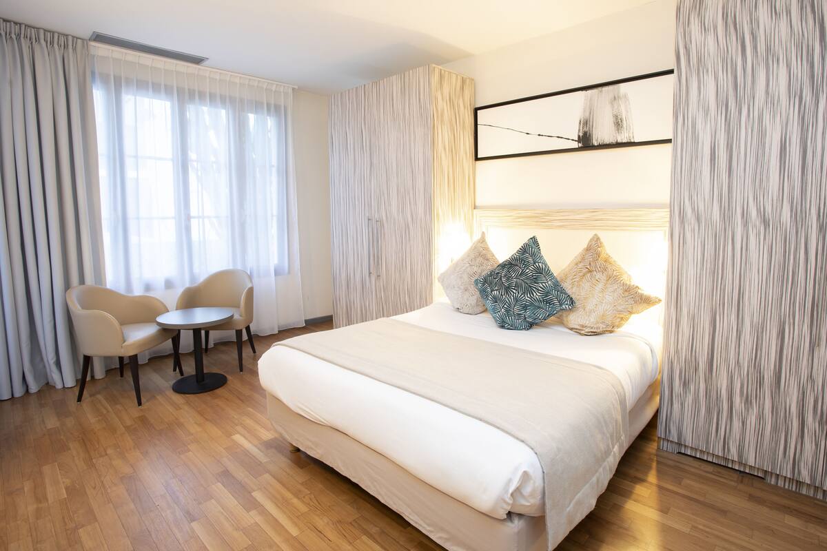 Une chambre d’hôtel avec un grand lit, une table ronde, deux chaises et une vue sur l’extérieur à travers des rideaux légers.